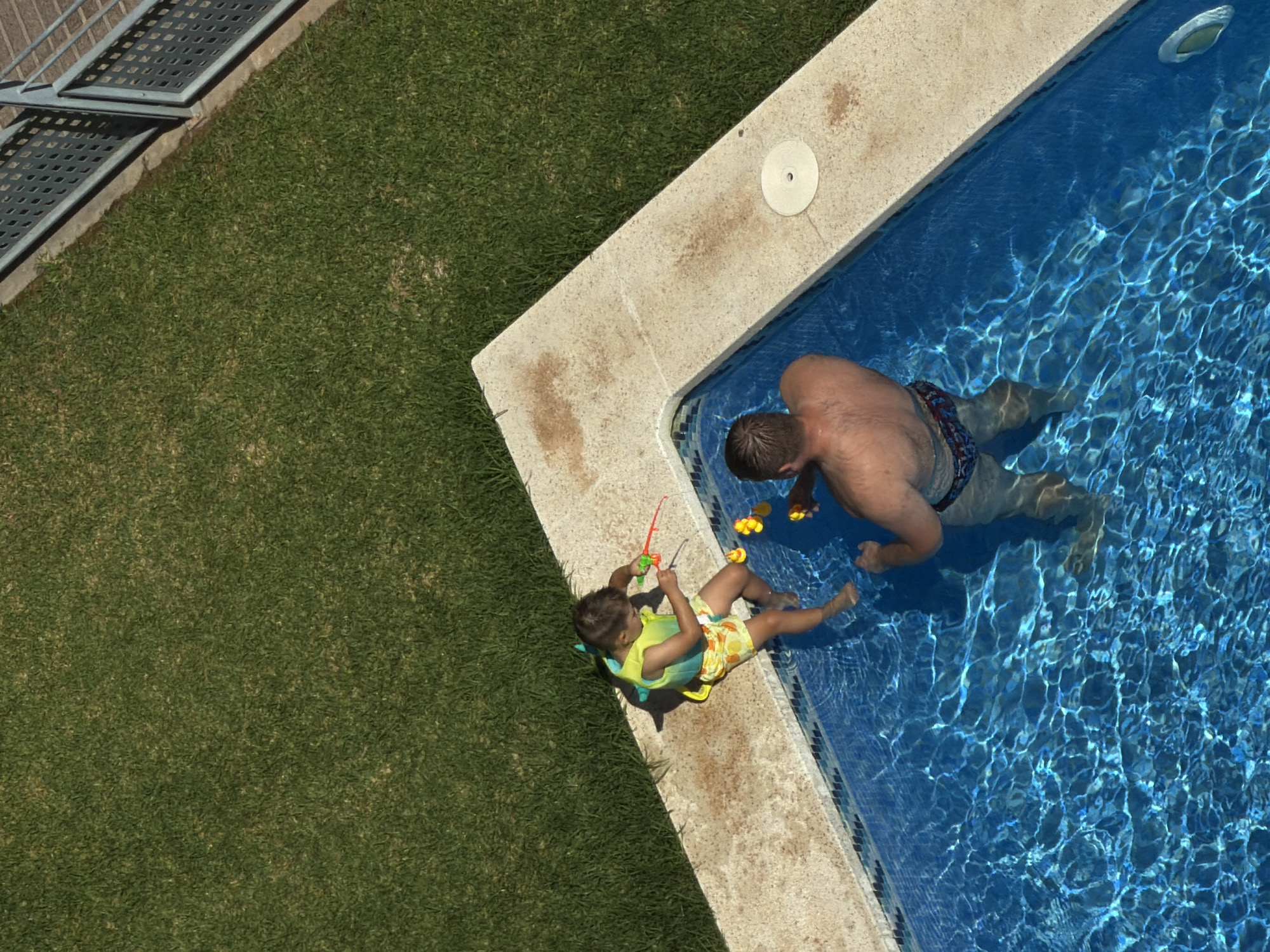 An aerial, high-angle shot captures a father and son engaged in a water gun fight by a swimming pool on a sunny day. The father, a man with short brown hair, stands waist-deep in the crystal-clear blue water, wearing patterned swim trunks. He is facing away from the camera, with his back and shoulders visible. To his left, on the pool's edge, sits the son, a young boy with dark hair, wearing a yellow and blue life vest and patterned swim trunks. He is holding a red and green water gun and appears to be aiming it. Yellow plastic ducks float in the pool near the boy. The pool's edge is made of beige concrete. Beyond the pool, a vibrant green lawn stretches out, meeting a metal fence at the far edge of the frame. The overall scene suggests a leisure activity in a residential or recreational outdoor setting.