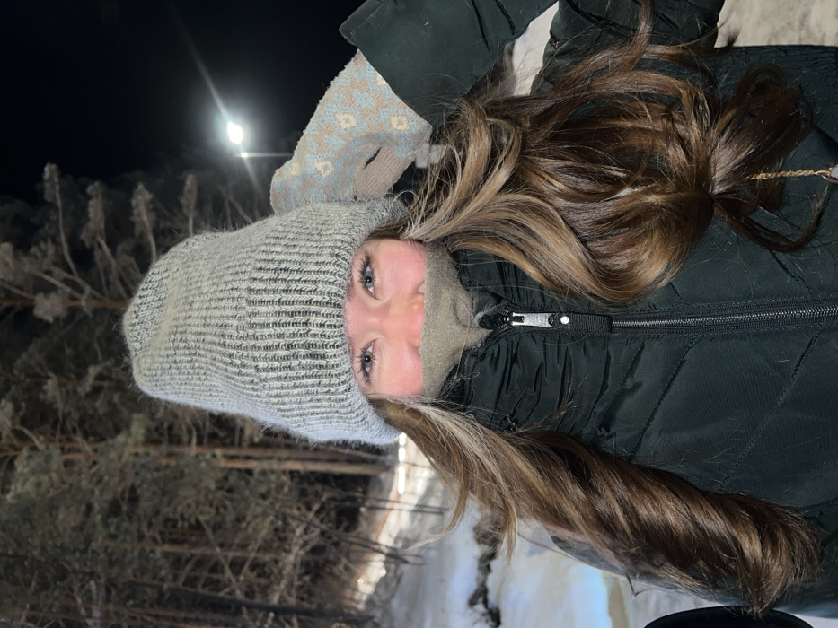 An individual is depicted outdoors at night in a snowy, forested environment near Krasnoyarsk, Russia. The person, positioned centrally and looking directly at the camera, appears to be smiling. They are wearing a light gray knitted beanie that covers their ears and forehead, a dark green puffer jacket zipped up, and a light olive-colored neck gaiter or scarf pulled up to cover their nose and mouth. Long, wavy brown hair with lighter highlights is visible cascading over their shoulders and down their chest. Their blue eyes are prominent, and their cheeks show a slight flush. The person's right hand, clad in a patterned light blue and beige mitten, is raised near the right side of their head, touching the beanie.
The background consists of snow-covered ground and numerous tall, slender conifer trees, also dusted with snow, indicating a cold, winter setting. The scene is illuminated by artificial light sources; a bright, tall streetlight is visible in the upper right portion of the image. The ground slopes downwards slightly from the foreground to the background. The overall impression is a candid shot taken in a cold, natural area at night.