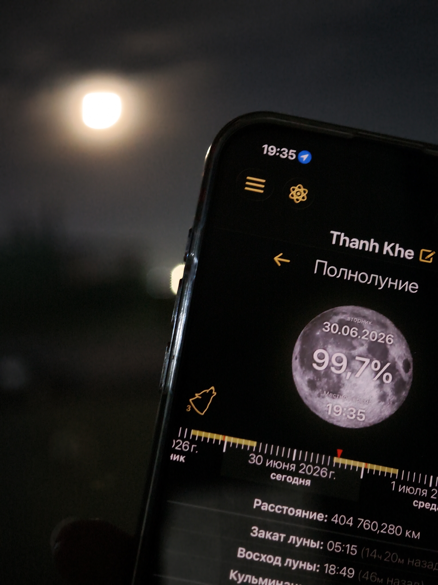A close-up shot captures a smartphone displaying a moon phase application against a dark, night sky. The phone's screen is the primary focus, showing detailed information about the moon. The date "30.06.2026" is prominently displayed, along with a percentage "99.7%" indicating a full moon. The local time is shown as "19:35". The screen also features a timeline with markings for "30 июня 2026 г. сегодня" (June 30, 2026, today) and "1 июля 2 среда" (July 1, Wednesday), suggesting the current date is June 30, 2026. Additional details visible include "Расстояние: 404 760,280 км" (Distance: 404,760,280 km), "Закат луны: 05:15" (Moonset: 05:15), and "Восход луны: 18:49" (Moonrise: 18:49). A stylized wolf graphic is also present on the screen. In the background, a bright, blurred light source, likely the full moon itself, illuminates the dark sky. The scene suggests the user is observing or tracking the full moon from Đà Nẵng, Vietnam, during the evening. The focus on the phone screen implies an interest in astronomy or the moon's cycle. The overall mood is one of nighttime observation and digital engagement with celestial events.