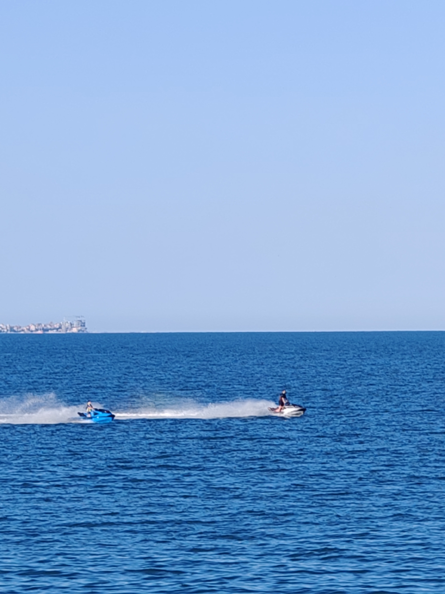 The image shows two jet skis speeding across the Black Sea near Burgas, Bulgaria. The sky is clear and blue, the sea is deep blue with visible wave patterns. On the left side of the frame, a blue jet ski is ridden by one person, leaving a long white trail of water. Further right, another jet ski, predominantly white and black, also with one rider, is moving in the same direction, similarly creating a white water trail. In the distant background, a low skyline with buildings is visible along the horizon.
