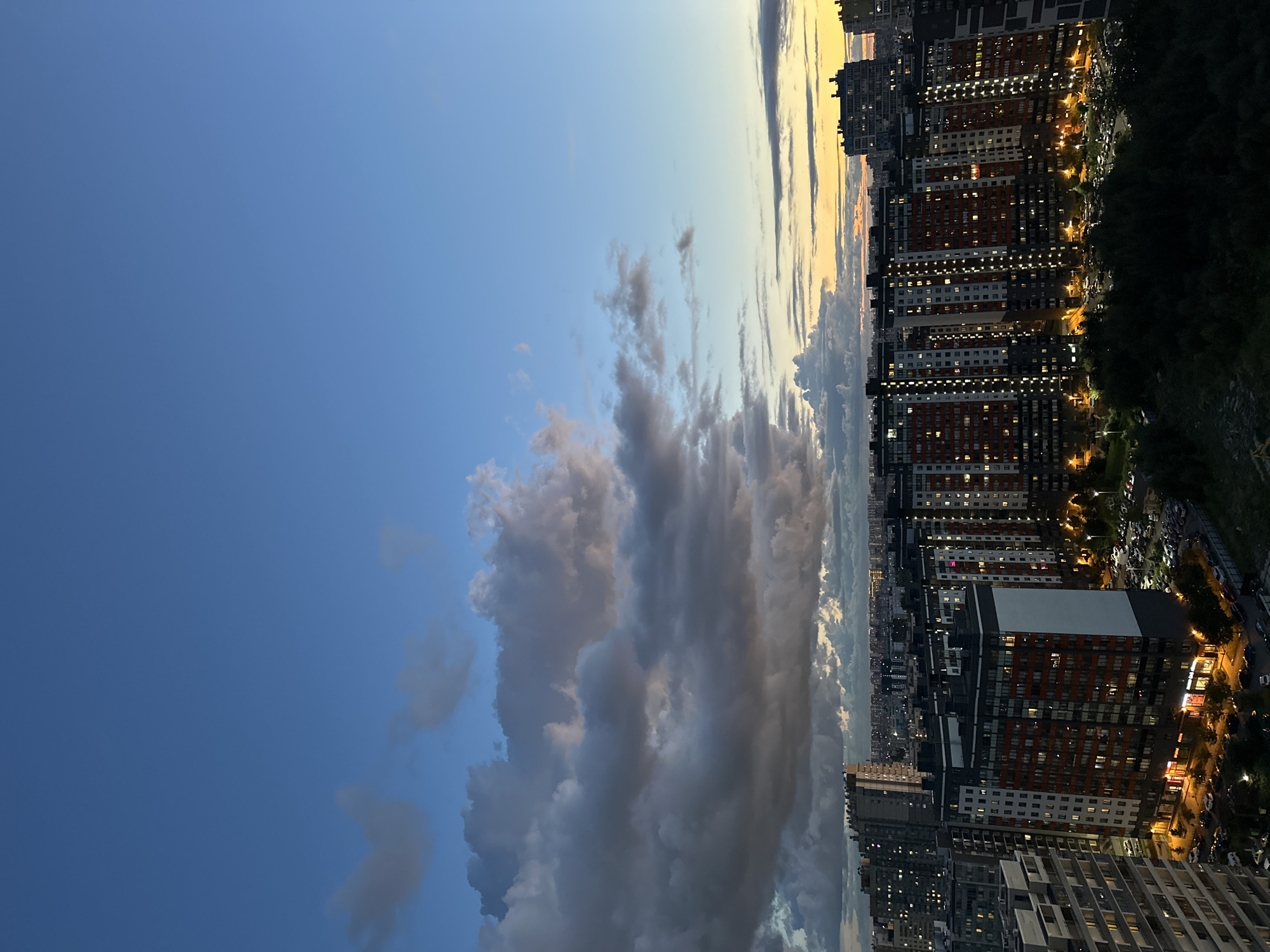 The image is a high-angle view of the city of Kudrovo, Russia, at dusk. The sky is a gradient of deep blue at the top, transitioning to lighter blues and then to a warm orange and yellow at the horizon, indicating sunset. Several large, dark clouds are present in the upper left portion of the sky, with some lighter clouds scattered elsewhere.
Below the sky is a dense urban landscape composed of numerous residential buildings. In the foreground, several tall, multi-story apartment buildings with modern architecture are prominent. These buildings have a dark gray base and facades with sections of red and white, and many of their windows are illuminated, suggesting that people are inside. The buildings are closely packed together, creating a sense of density.
In front of the buildings, there are parking lots filled with numerous cars. Trees and greenery are visible in the lower portion of the image, especially to the right, appearing dark against the illuminated buildings and sky. Further in the distance, more buildings stretch towards the horizon, forming a sprawling cityscape under the twilight sky. There are no visible people or animals in the image. The overall scene depicts a residential area of a city during the transition from day to night.