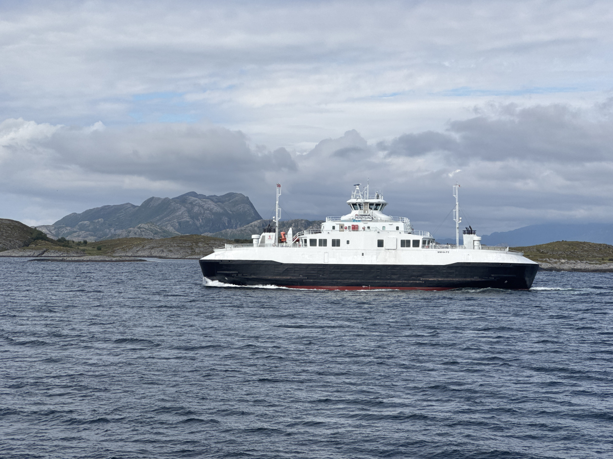 A large ferry, identified by the text "MM64FD" and "HERØYSUND" on its superstructure, travels through choppy, dark blue water. The ferry has a white upper deck and a black hull with a red waterline. It is moving from right to left in the frame. In the background, rocky, green-brown islands and mountains are visible under a cloudy, overcast sky. The scene suggests a somewhat cool and potentially breezy day. The ferry appears to be in motion, creating a wake behind it. The location is likely Stokkvågen, Norway, given the reference provided. There are no people clearly visible on the deck of the ferry, and the overall mood is neutral, focusing on the maritime landscape and the vessel.