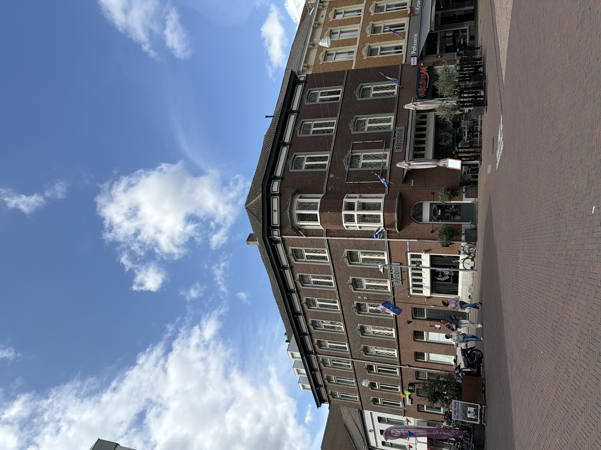 The image depicts a street scene in Roermond, Netherlands, with a focus on the facade of a brick building housing a restaurant named "RHODOS". The building has multiple floors with numerous windows, many of which are ornate. Flags, one with a blue and white pattern, are visible on the building. In front of the restaurant, tables and chairs are set up outdoors under umbrellas, suggesting a cafe or dining area. Adjacent to "RHODOS" are other businesses, including "Pâtisserie" and "de Kroon". Several people are walking on the brick-paved sidewalk in front of the buildings, and a scooter and bicycles are parked nearby. The sky above is blue with scattered clouds. The overall scene suggests a moderately busy urban environment on a pleasant day.