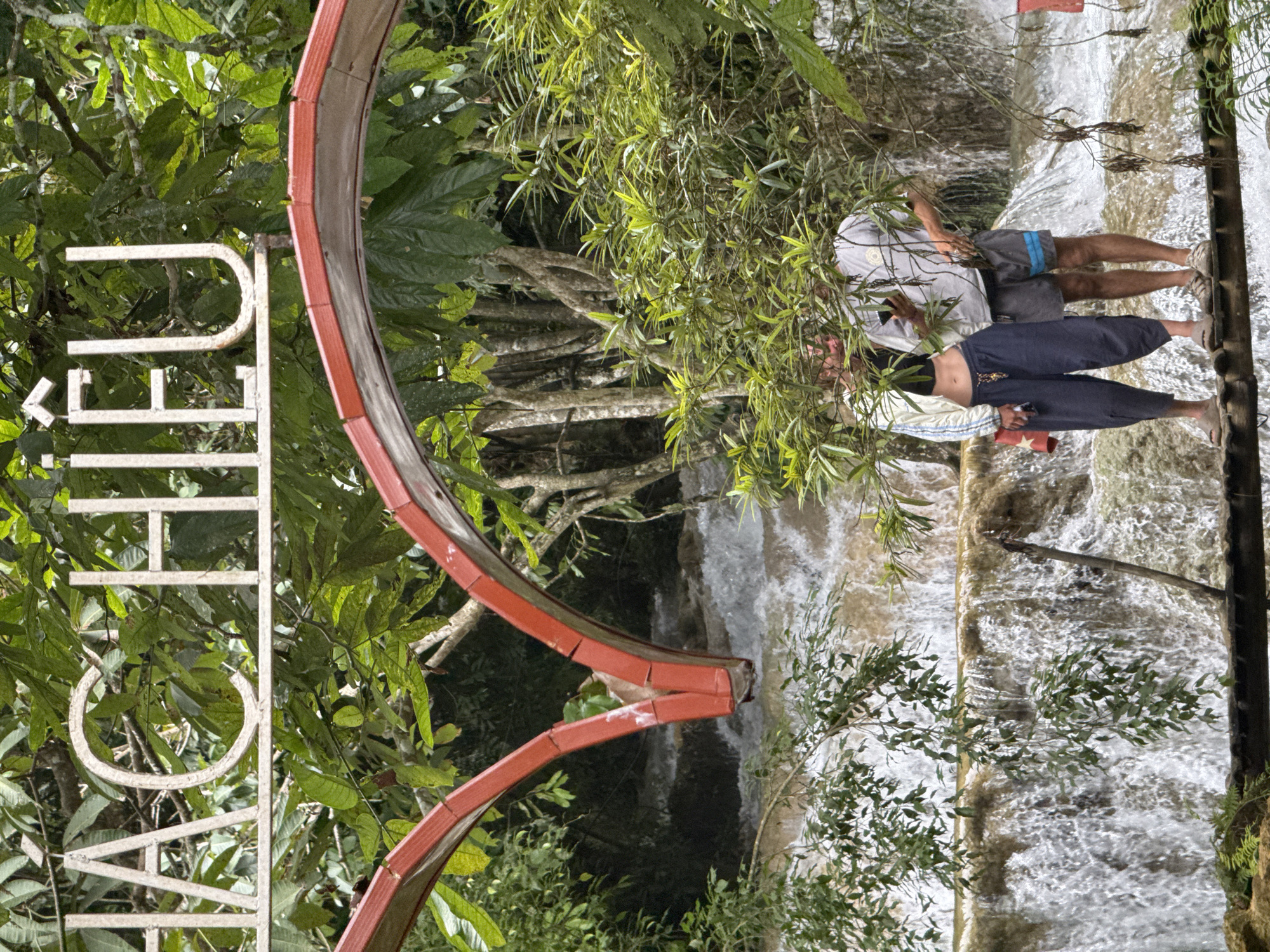 The image shows a waterfall in Cổ Lũng Commune, Vietnam, with a sign overhead that reads "HÀ CHIÊU". The sign is white metal with a red heart-shaped arch below it. In the foreground, two people are standing on a wooden bridge, looking at a cell phone. The woman is wearing a white jacket, a black crop top, and blue pants. The man is wearing a gray t-shirt and gray shorts. The waterfall is brown and turbulent, suggesting recent rainfall. The surrounding environment is lush and green with trees and foliage.