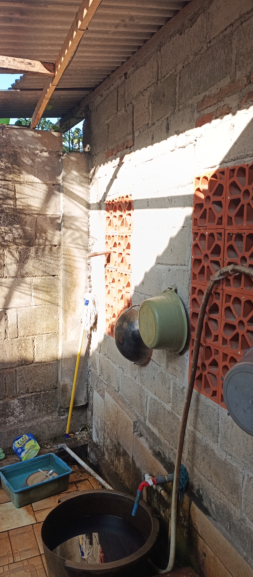 In an outdoor utility area, a grey cinder block wall is visible, partially covered by a corrugated metal roof. Two decorative orange brick inserts are set into the wall. A green plastic bucket and a dark basin are hanging on the wall, with a brown hose coiled nearby. In the foreground, a large, dark plastic tub filled with water sits on a tiled floor. A blue plastic basin with dishes inside is next to the tub, and a yellow mop with a white head is propped against the wall. A package of "KILAU NIPIS" is also on the floor. A red and white faucet is attached to a pipe on the wall. Sunlight casts shadows across the scene.