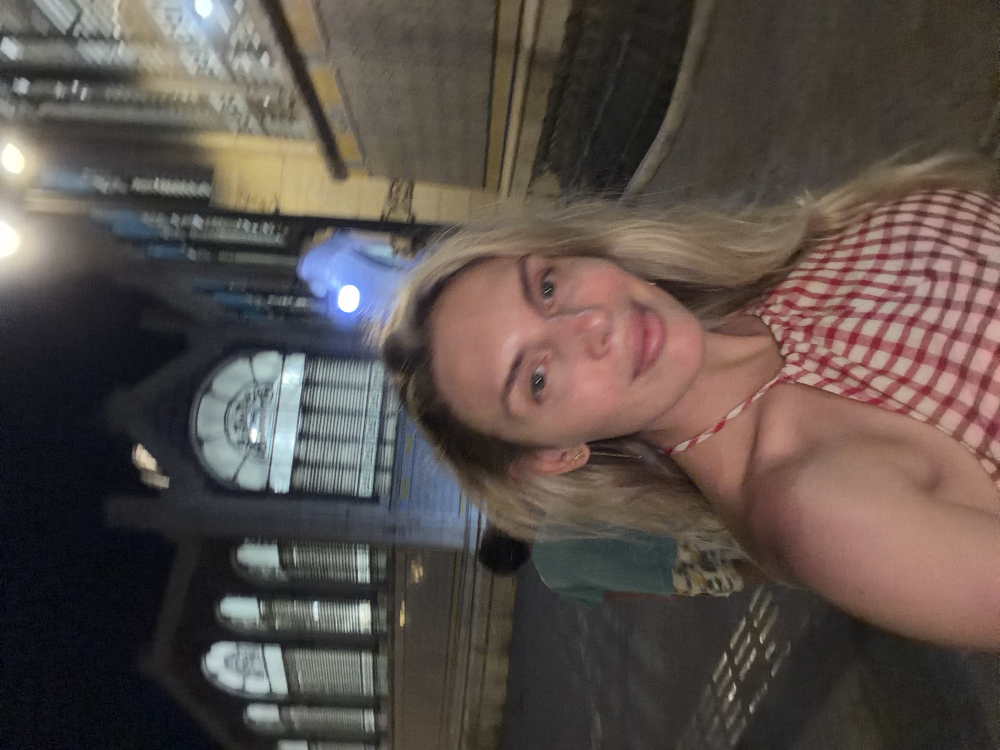 A woman with blonde hair, wearing a red and white checkered top, is taking a selfie at night in a city. In the background, a young person wearing a teal t-shirt and patterned shorts is walking away. Behind them, a large, arched building with striped windows is illuminated. The street appears to be paved, and there are streetlights casting shadows. The scene suggests a public urban space, possibly a plaza or a street in Valencia, Spain, given the architectural style.