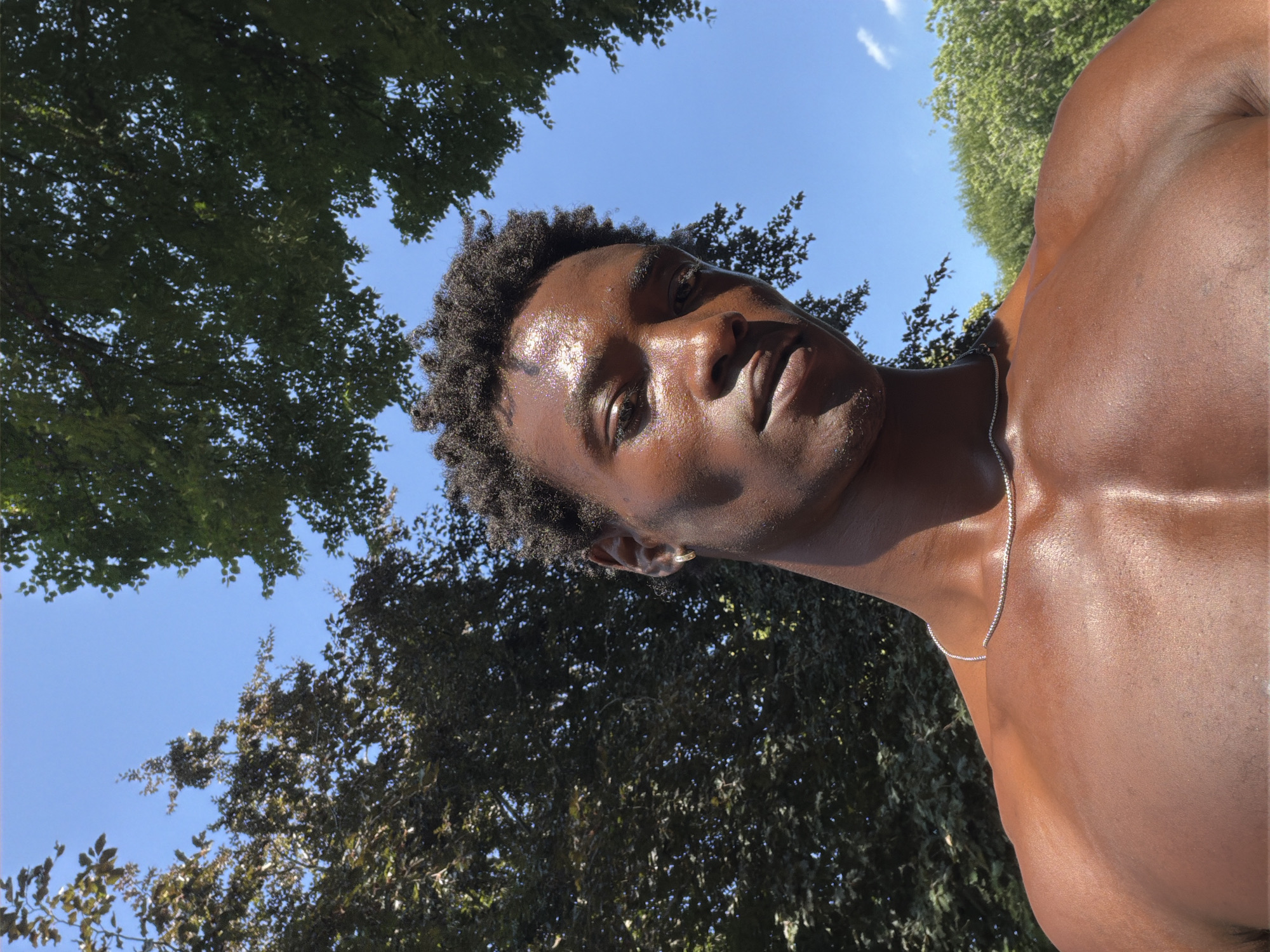 A shirtless man with dark skin and short, curly dark hair looks up at the camera. He has sweat glistening on his face and chest. He is wearing a thin silver chain necklace. The man is outdoors, with a bright blue sky visible above him and the tops of green and dark brown trees framing the shot. The image appears to be taken from a low angle, looking up at the man. The background suggests a park or natural area. There is no indication of location in the city of London, United Kingdom.