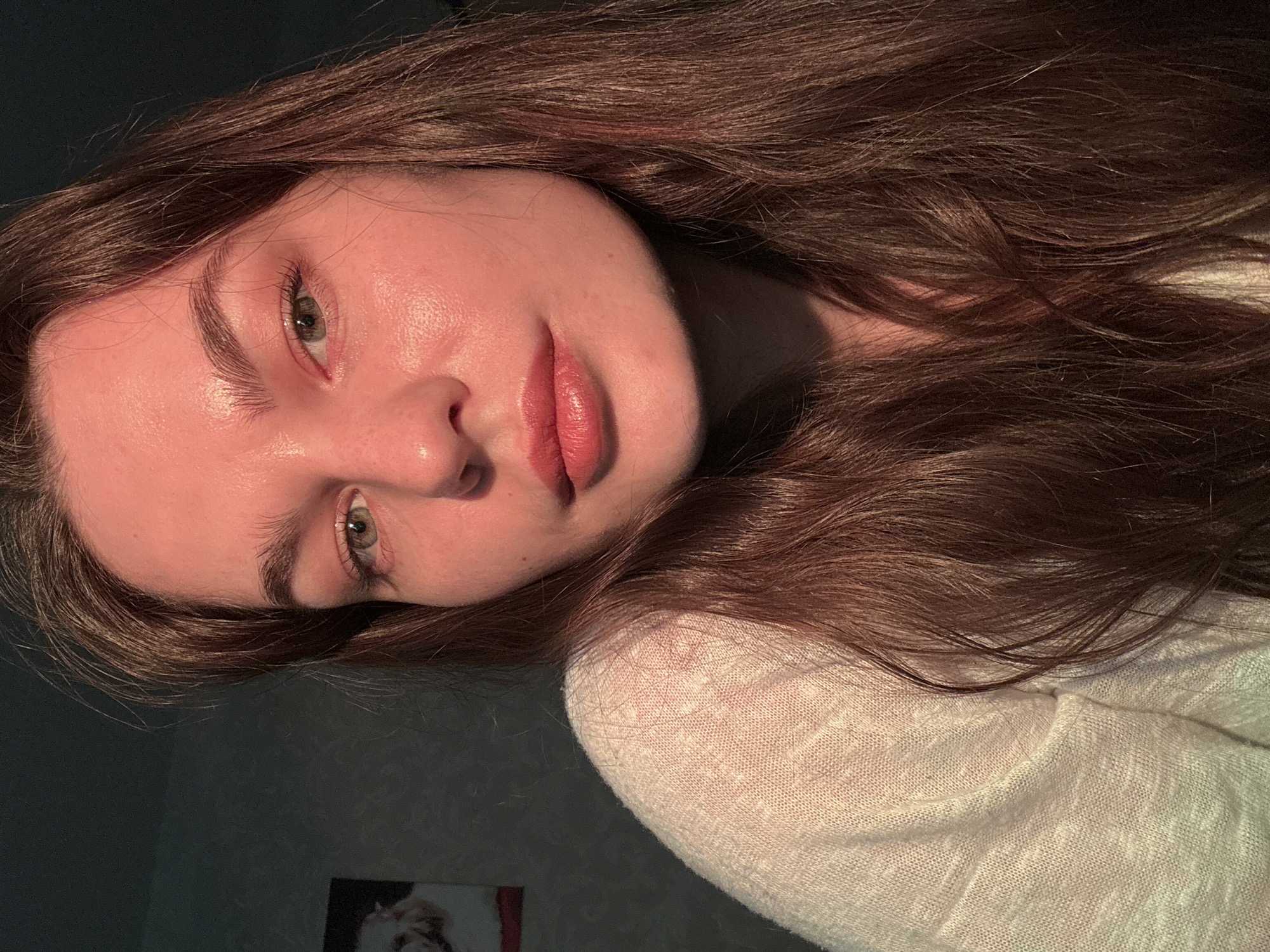 A medium shot, chest-up, selfie-style portrait shows a young woman with long, dark brown hair. Her hair is parted in the middle and falls around her face and shoulders. She is wearing a light-colored, textured knit sweater. The lighting in the image is warm and directional, casting a reddish-orange glow on her skin, particularly on her face. Her left eye is visible, appearing to be green or hazel. Her lips are slightly parted and have a subtle rosy hue. The background is dark and appears to be an indoor setting, with a textured wall and a framed picture on the left side. The overall impression is that of a candid, natural shot taken in the evening or with artificial lighting. The location is specified as the city of Chaikovsky, Russia.
