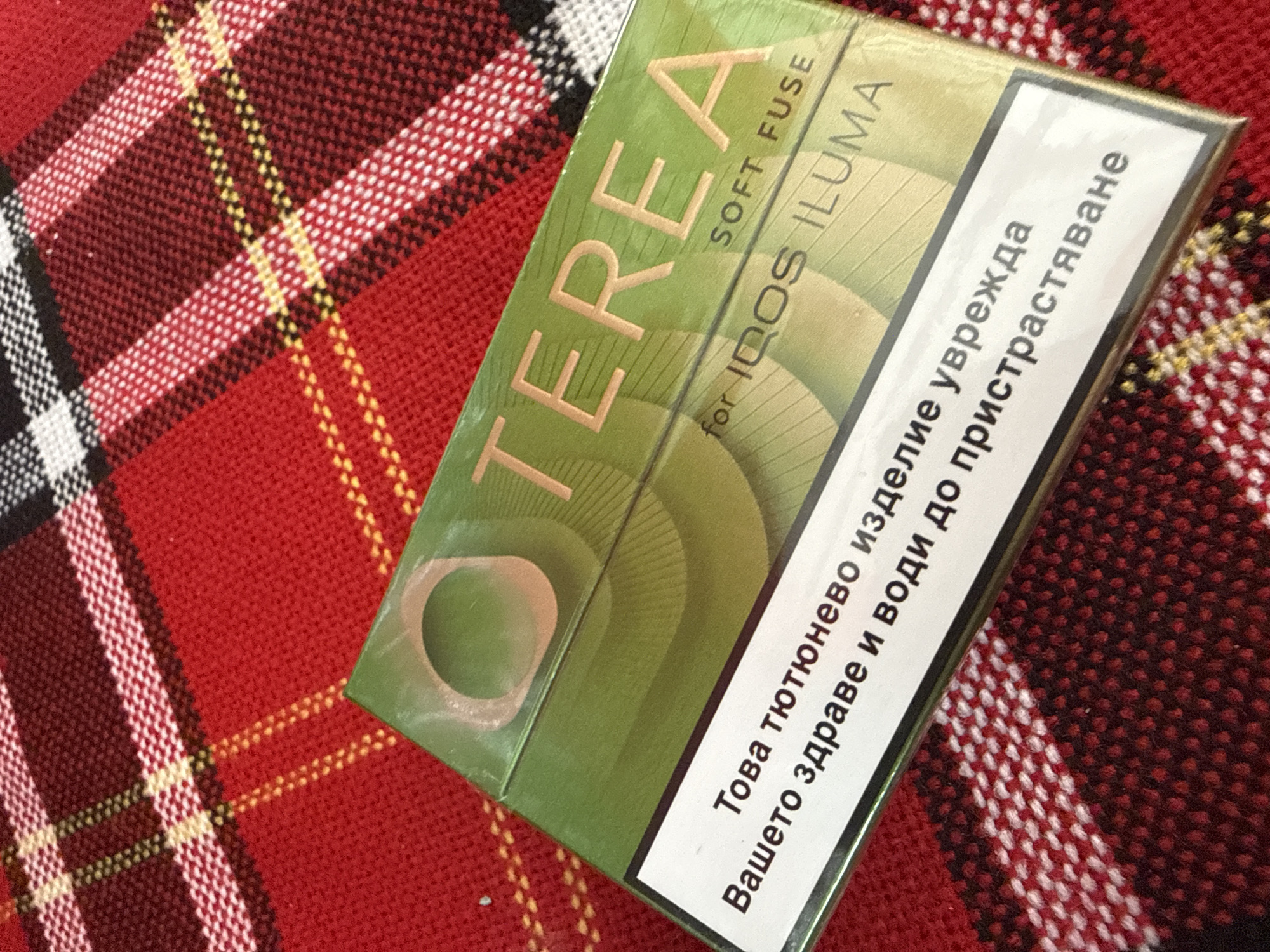 A top-down, medium shot shows a green and gold colored pack of "TEREA SOFT FUSE for IQOS ILUMA" tobacco sticks, positioned diagonally across a red and black checkered textile surface. The textile appears to be a blanket or tablecloth. The pack has visible text in a lighter color, and a white label with text in Bulgarian. The lighting is even, and the focus is sharp on the product. The scene is devoid of any human presence or discernible location markers that would indicate Sofia, Bulgaria.
