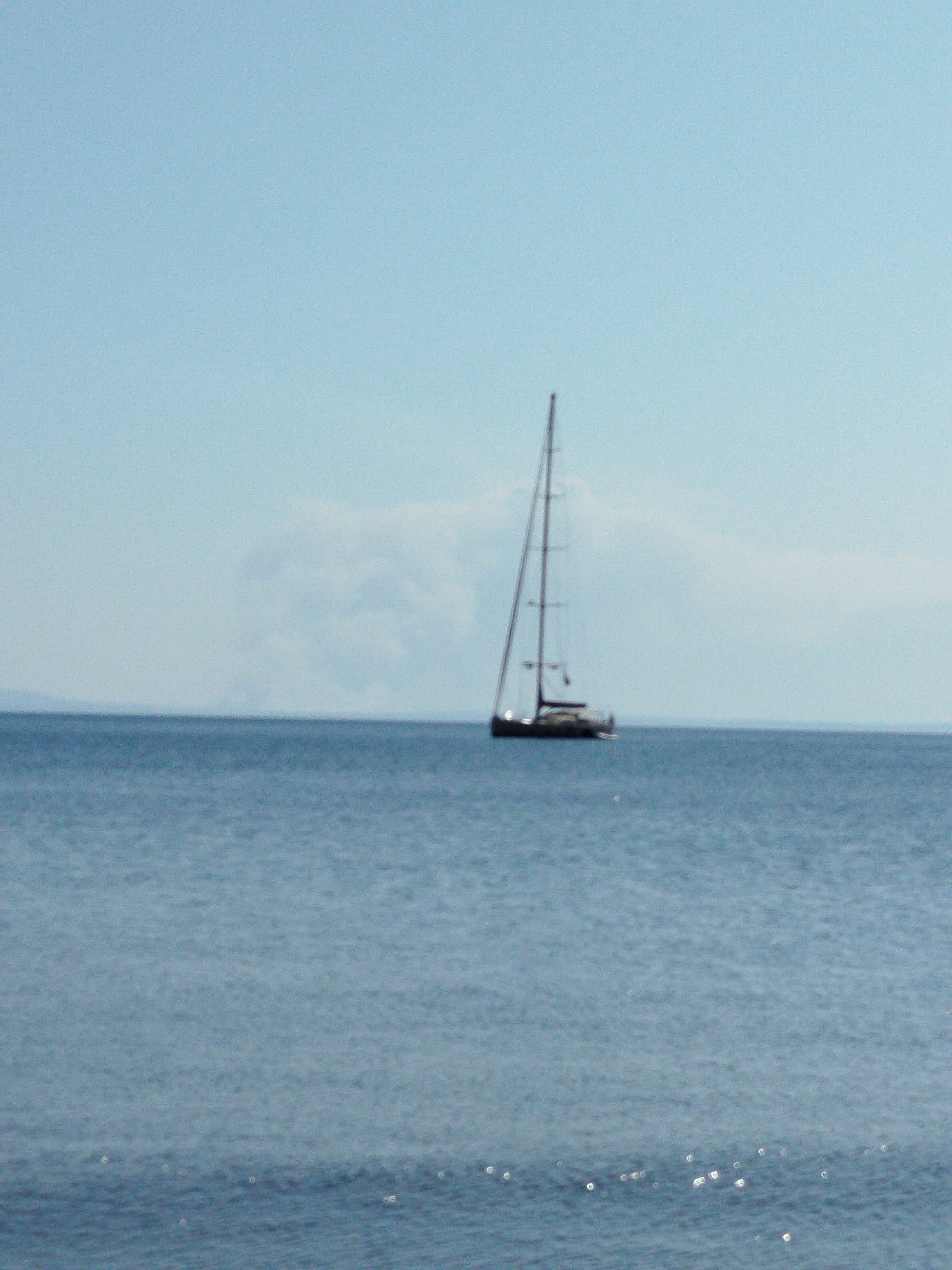 A sailboat with a tall mast is sailing on a calm blue sea under a clear, light blue sky. In the distance, a faint, hazy landmass is visible on the horizon, possibly mountains. The water has a slight ripple and some small glints of light on its surface. The image is shot from a low angle, looking across the water towards the sailboat. The setting is likely coastal, within the Sithonia Municipal Unit of Greece, given the characteristic clear blue waters and coastal scenery. There are no people visible in the image.