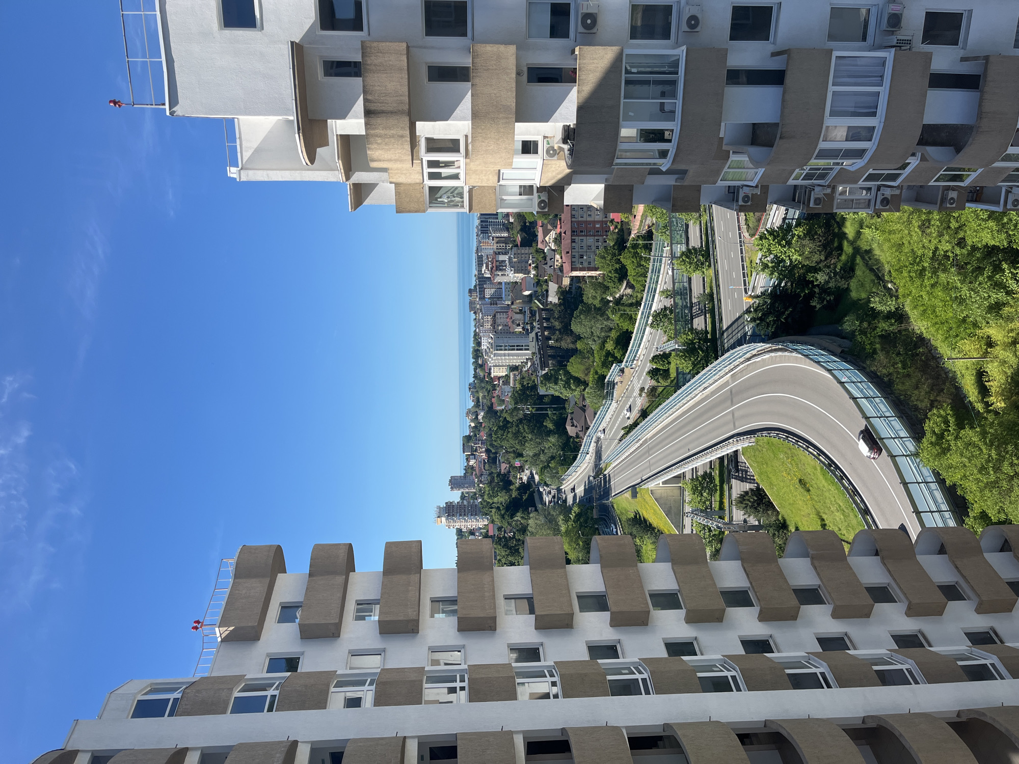 This vertically oriented image presents a high-angle view over a modern coastal city, likely Sochi, Russia. The scene is framed on the left and right by sections of two multi-story buildings, characterized by white facades and distinctive horizontal bands of brown, curved balcony or window awnings. Numerous rectangular windows and external air conditioning units are visible on these structures. A small red object, possibly a flag or warning device, is seen on a metal scaffold on the upper left building.
The central part of the image reveals a dynamic urban landscape under a clear blue sky with a few wispy clouds. A prominent feature is a multi-lane highway, winding through lush green hills and valleys. The road has clear white lane markings and is bordered by extensive greenery and a transparent or glass sound barrier. A white car is visible driving on a curved section of this highway. Beyond the green infrastructure, a dense cityscape stretches towards the horizon, filled with various buildings. The distant horizon features a vast expanse of blue water, indicative of the Black Sea, merging with the sky.
The bright sunlight suggests midday or early afternoon conditions. The perspective is from a high vantage point, offering a broad overview of the city's blend of modern infrastructure, urban development, and natural coastal beauty. No discernable text is visible in the image.