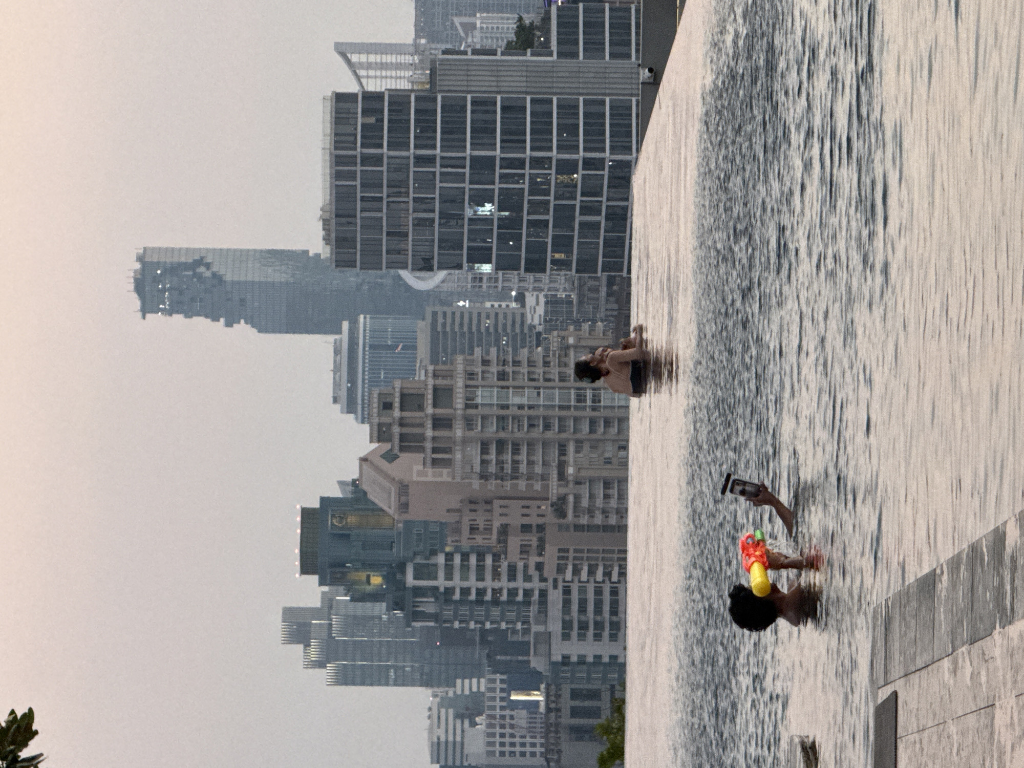 A sideways, eye-level shot shows two people enjoying an infinity pool overlooking the skyline of Bangkok, Thailand. The city's buildings are visible behind the pool, receding into the hazy atmosphere. The person closest to the camera has dark hair and holds a colorful water gun and a mobile phone. The other person is farther away in the pool. The pool water is light and reflective, with visible ripples and waves. The image is well-lit, but the city behind the pool is shrouded in a slight haze, obscuring details.