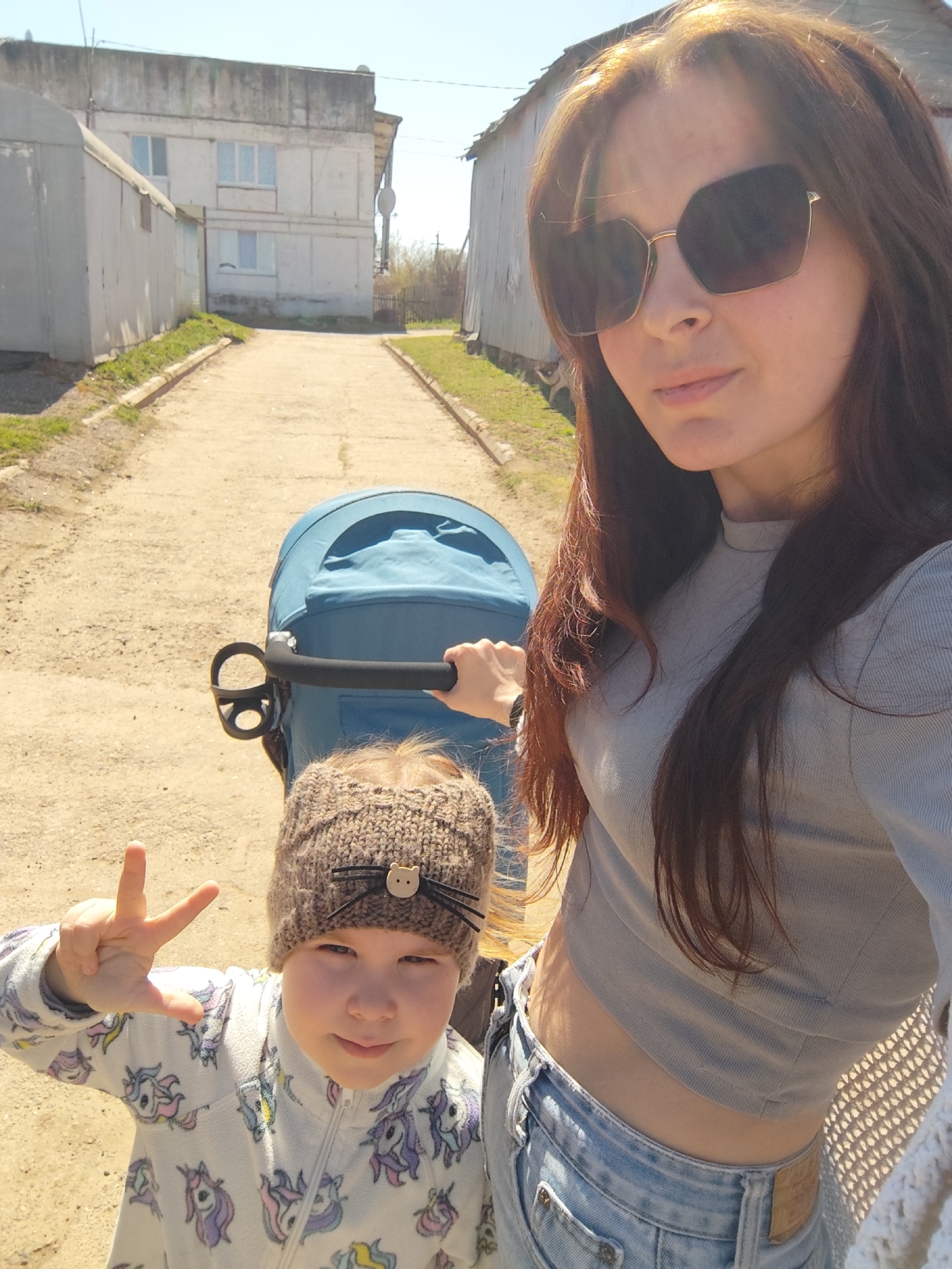 A woman with long brown hair and sunglasses stands on a paved path in Фоки, Russia. She is holding the handle of a blue stroller containing a child wearing a knitted hat with a cat face. The child is making a hand gesture. The woman is wearing a grey crop top and jeans. Buildings line both sides of the path, and the sky is clear.