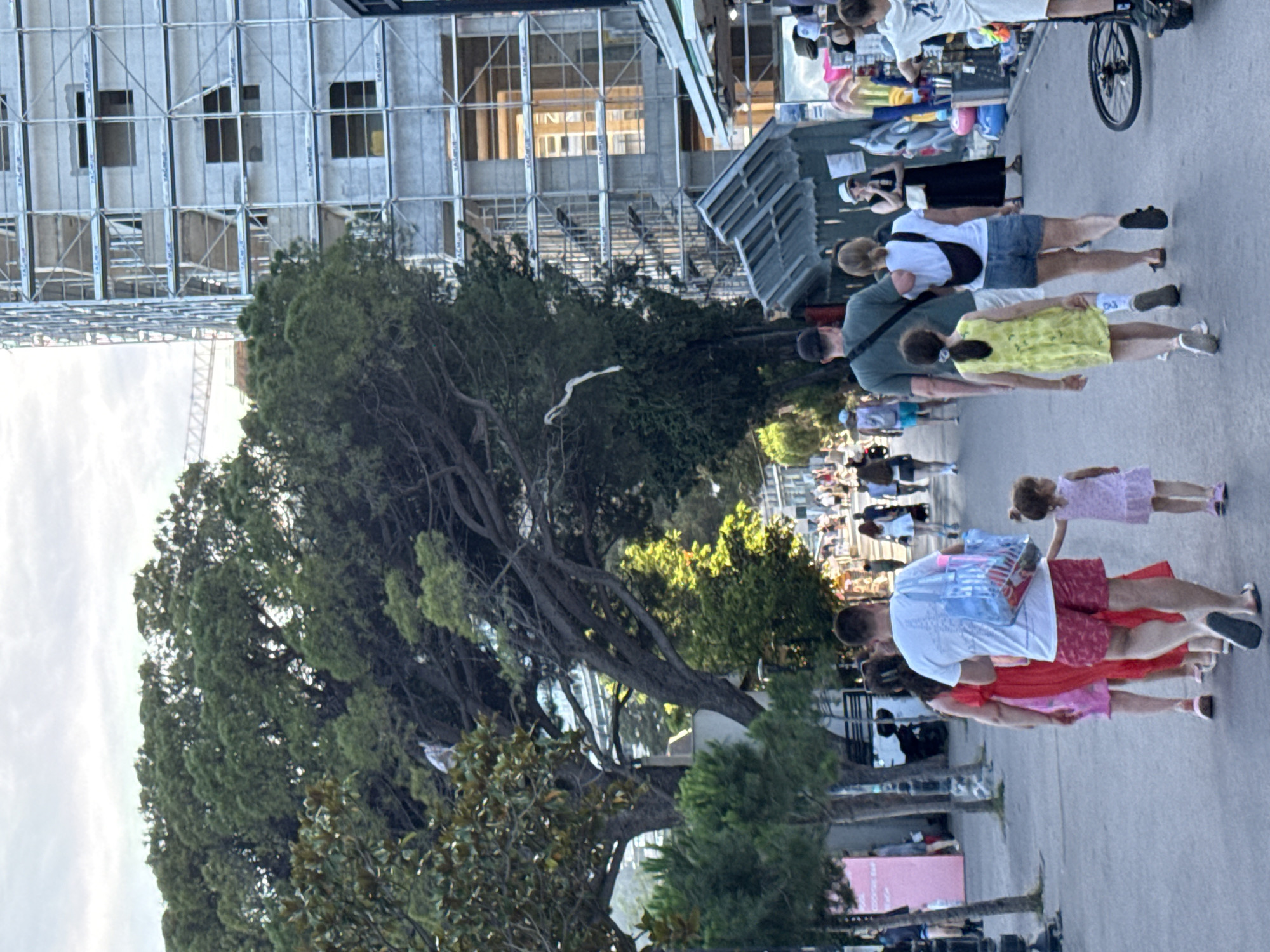This image captures a bustling pedestrian promenade in Budva, Montenegro, likely along the coastal stretch. The scene unfolds under an overcast sky, suggesting late afternoon or early evening.
In the foreground, a diverse group of pedestrians walks away from the camera. A man in a white t-shirt and red patterned shorts holds a clear blue plastic bag filled with items and is holding the hand of a small girl in a light pink dress. To his left, a woman in a pink dress walks beside him, and another woman in a white t-shirt, denim shorts, and a black fanny pack walks alongside a young girl in a yellow dress. A black bicycle lies on its side to the right.
The midground features a wide asphalt path flanked by dense, mature trees, predominantly conifers on the left and broadleaf trees on the right. On the right side of the path, several open-fronted market stalls or kiosks are visible, displaying colorful merchandise such as inflatable toys and hats. A woman in a black dress and white hat stands near one of these stalls. On the left, a partially visible pink sign advertises a "COCKTAIL BAR."
In the background, a large, multi-story building is under active construction on the right, completely enveloped in silver metal scaffolding. The brand name "TALMUR" is distinctly visible on multiple sections of the scaffolding. The building appears to be concrete with numerous square window openings. Beyond the trees on the left, parts of other structures are faintly visible, and a large white construction crane is discernable in the distant background on the left.