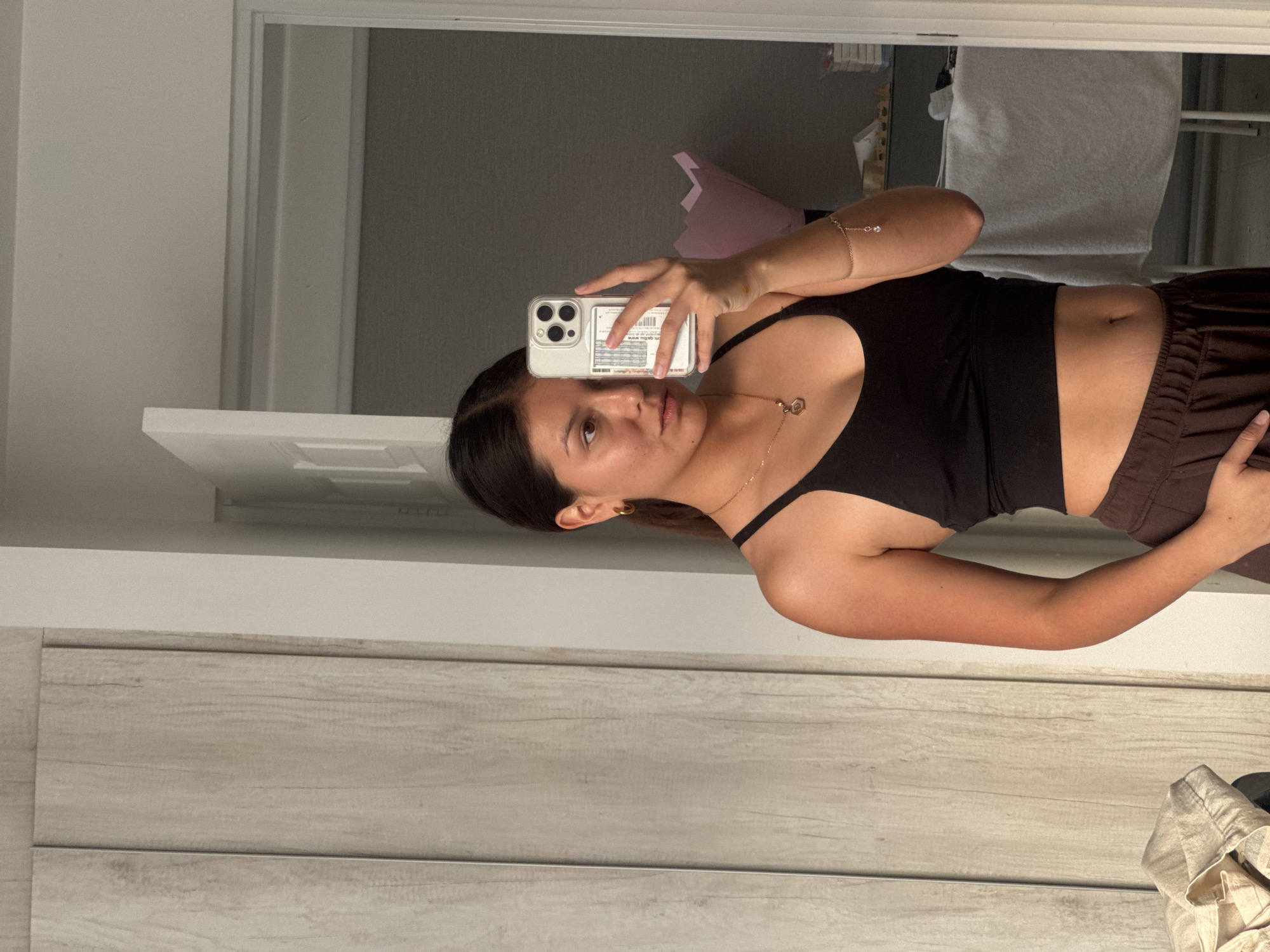 A person stands indoors and takes a picture of themself in a mirror with a smartphone. The individual has dark hair and brown eyes, wears a black crop top, and brown pants. The mirror shows a room with furniture and white walls. A white door frame is on the left. The floor has a wood grain pattern.