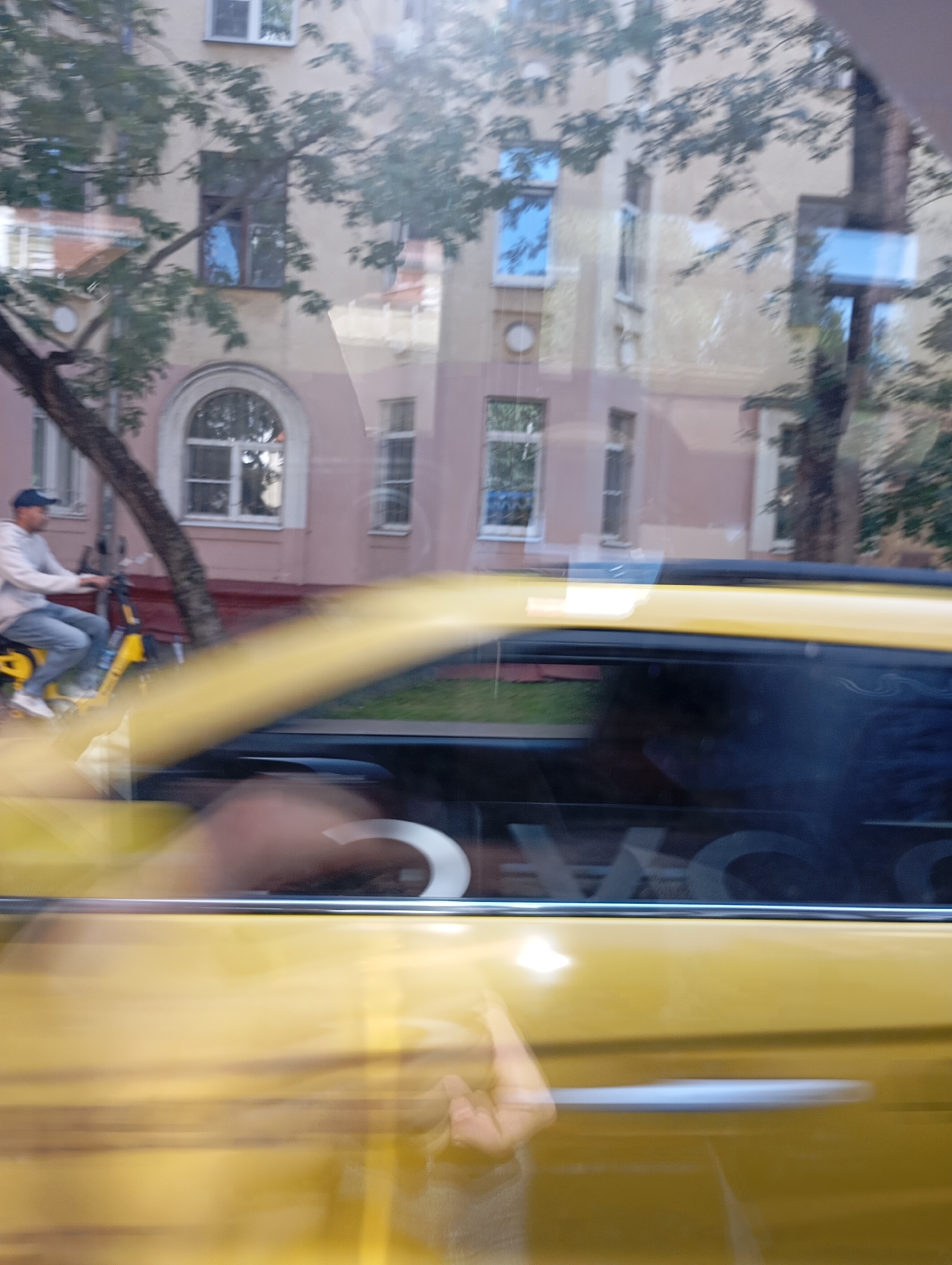 The image captures a street scene in Moscow, Russia, from the perspective of a moving vehicle. A person is visible riding a yellow electric scooter in the background, wearing a blue cap, light-colored sweatshirt, and jeans. Behind the rider, a multi-story building with pink and beige facades and numerous windows is reflected in the glass. A tree with green leaves partially obscures the building. In the foreground, the yellow exterior of the vehicle the image is taken from is prominent, with a blurred hand visible reaching towards the camera. The overall impression is one of motion and urban life, with a strong emphasis on reflections and the blur of speed.