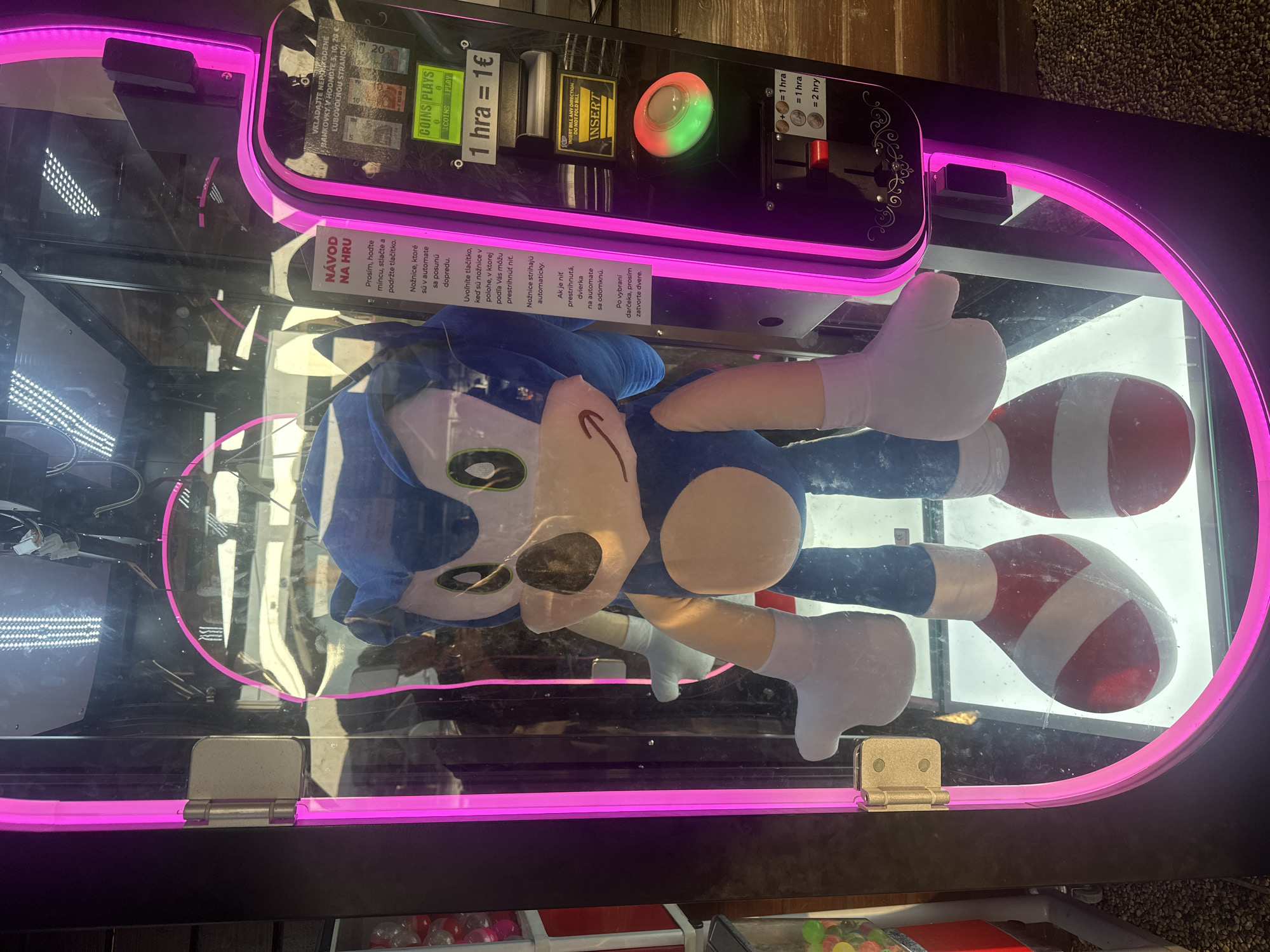 A close-up, eye-level shot captures a Sonic the Hedgehog plush toy inside a brightly lit arcade claw machine. The machine, adorned with pink neon lights, stands in what appears to be an indoor establishment, possibly a game arcade or a shopping mall, in Zamarovce, Slovakia. The plush toy, the main subject, is positioned centrally, its blue fur, iconic red and white shoes, and white gloves are clearly visible. The machine's interface is on the right, featuring a coin slot, buttons, and instructions in Slovak. A sign above the coin slot reads "NÁVOD NA HRU" (Instructions for Play). The machine is designed to allow players to attempt to win prizes, like the plush toy, by operating a mechanical claw. The background is a blurred reflection of the surroundings, suggesting a public space with people and other machines.
