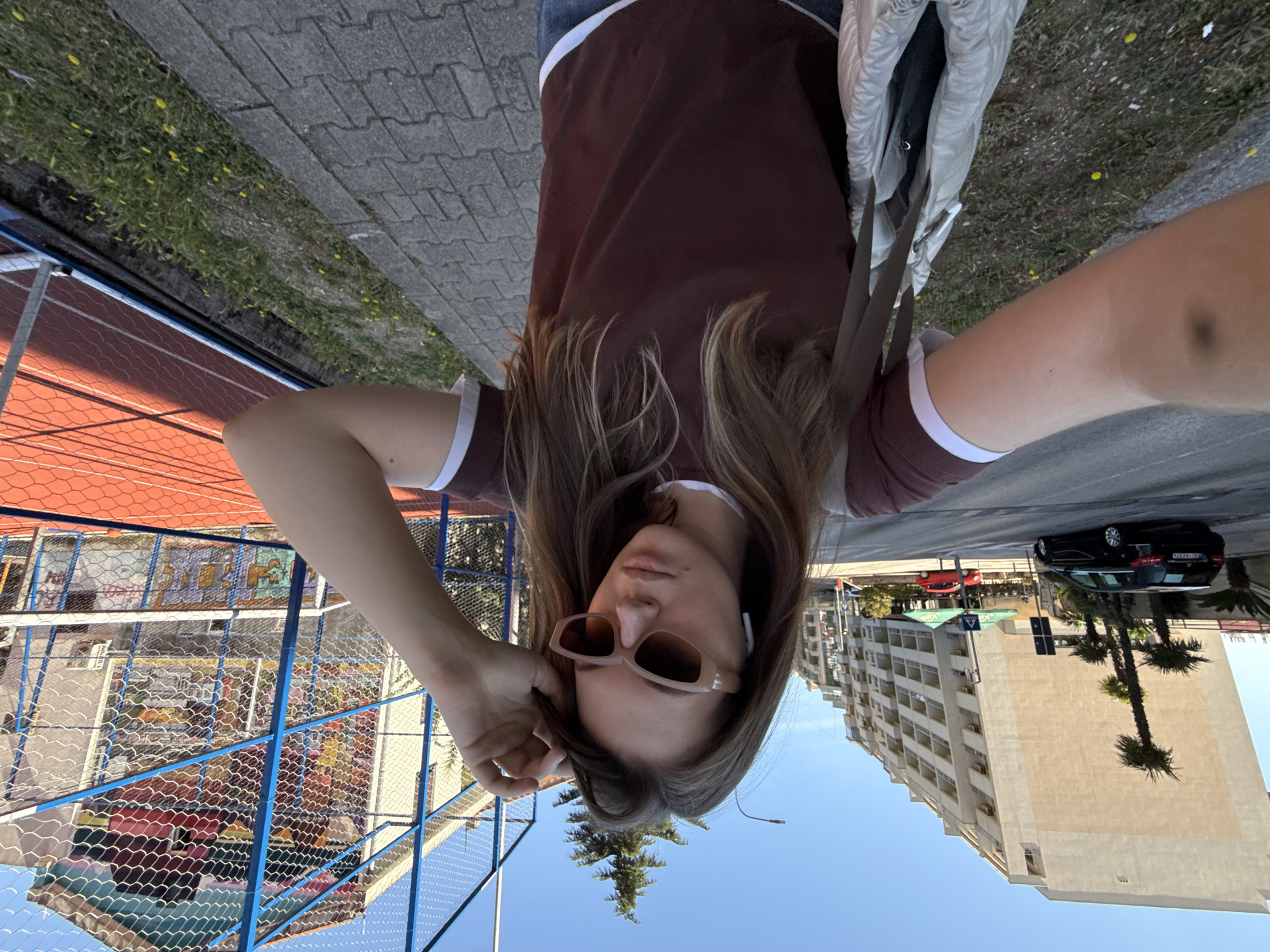 The image displays a young adult female taking a selfie on a sidewalk in Budva, Montenegro, under a clear blue sky. She has long brown hair, wears light-colored cat-eye sunglasses, and white wireless earbuds. Her left hand is raised, touching her hair. She is dressed in a dark reddish-brown t-shirt with white trim on the collar and sleeves, blue jeans, and carries a light-colored drawstring shoulder bag on her left side. A subtle dark mark is visible on her right upper arm.
To her left, an asphalt road features a black Mercedes-Benz car in the mid-ground and a red compact car further in the distance. Several palm trees line the road. Beyond the road, multiple multi-story residential buildings with balconies, light-colored facades, and visible air conditioning units are present, with one building on the far left exhibiting a prominent angled wall.
To her right, a narrow grassy strip containing small yellow flowers separates the paved sidewalk from a red-surfaced sports court. The court is enclosed by a blue-framed white mesh fence, with a net visible, consistent with a tennis court. Behind the court, a building with colorful murals on its exterior walls is partially observable through the fence.