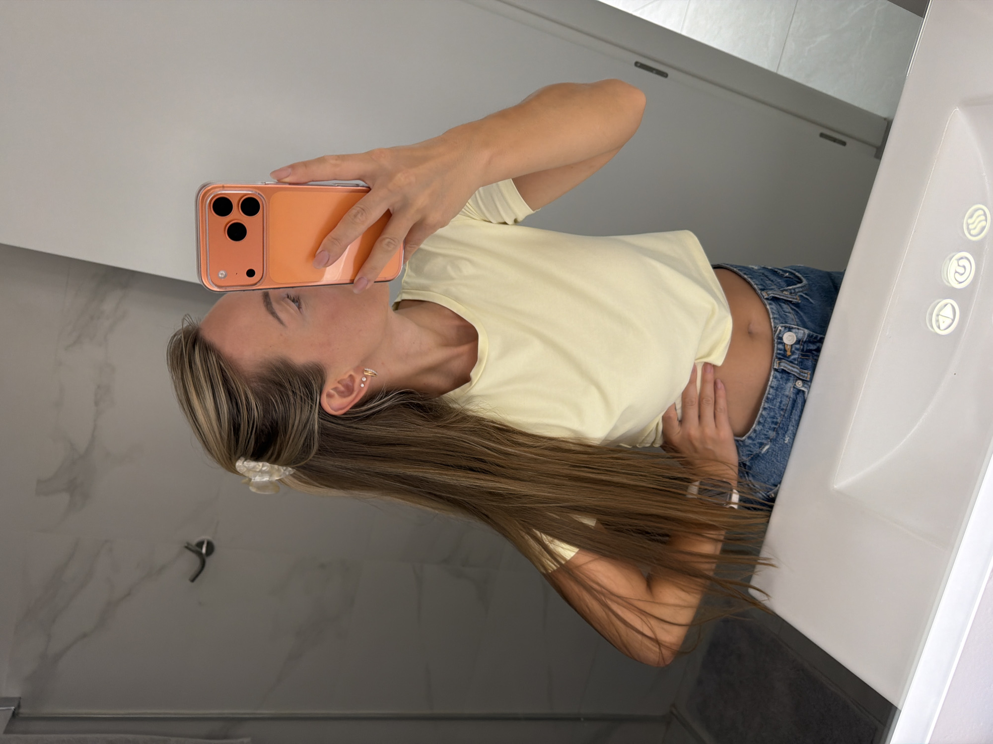 In a well-lit bathroom, a woman with long, blonde, highlighted hair pulled back with a white claw clip takes a mirror selfie. She is wearing a light yellow, cropped t-shirt and blue denim jeans. Her left hand rests on her hip, and her right hand holds an orange smartphone to capture the image. She is wearing small hoop earrings. The bathroom features a marble-tiled wall behind her and a white sink in the foreground. The scene appears to be in Finestrat, Spain, a coastal town known for its scenic beauty.
