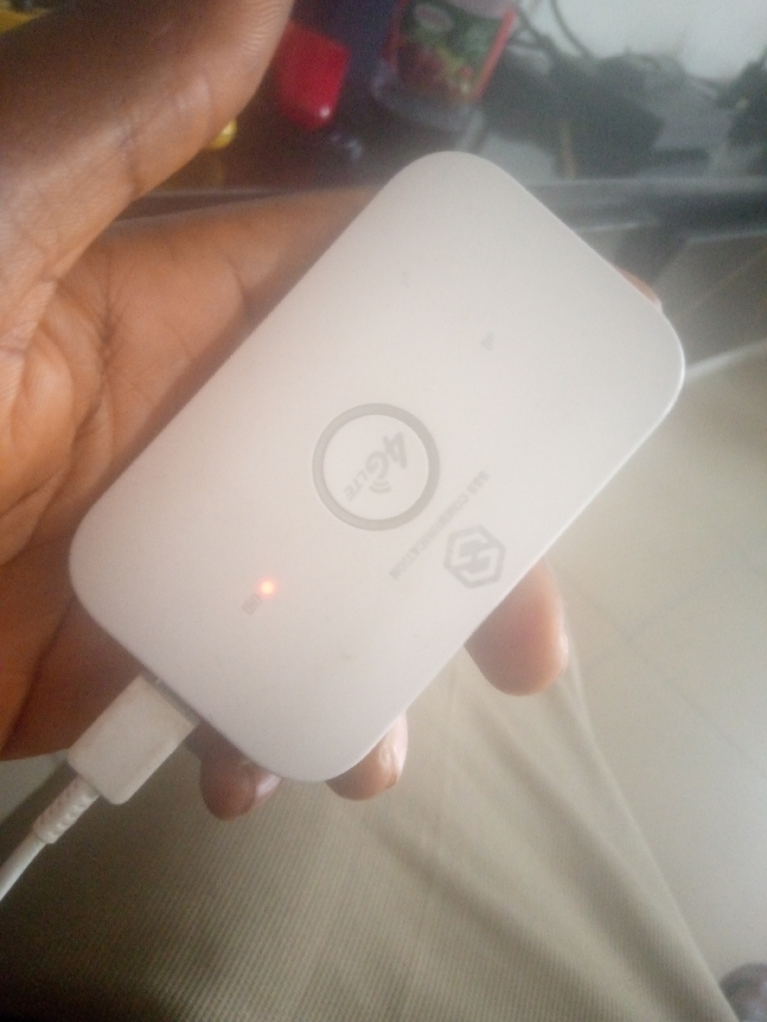 In a close-up, indoor shot, a person's hand, with dark skin and visible fingerprints, is holding a white portable Wi-Fi hotspot. The hotspot has a circular "4G LTE" logo and other subtle markings on its surface. A small red LED light is illuminated on the device, indicating it is active or charging. A white USB cable is plugged into the bottom of the hotspot and extends out of frame. The background is blurred, suggesting an out-of-focus environment, but faint shapes of objects and colors, possibly in a room, are visible. The scene is not definitively identifiable as being in Jalingo, Nigeria, as no specific geographic or cultural markers are present.