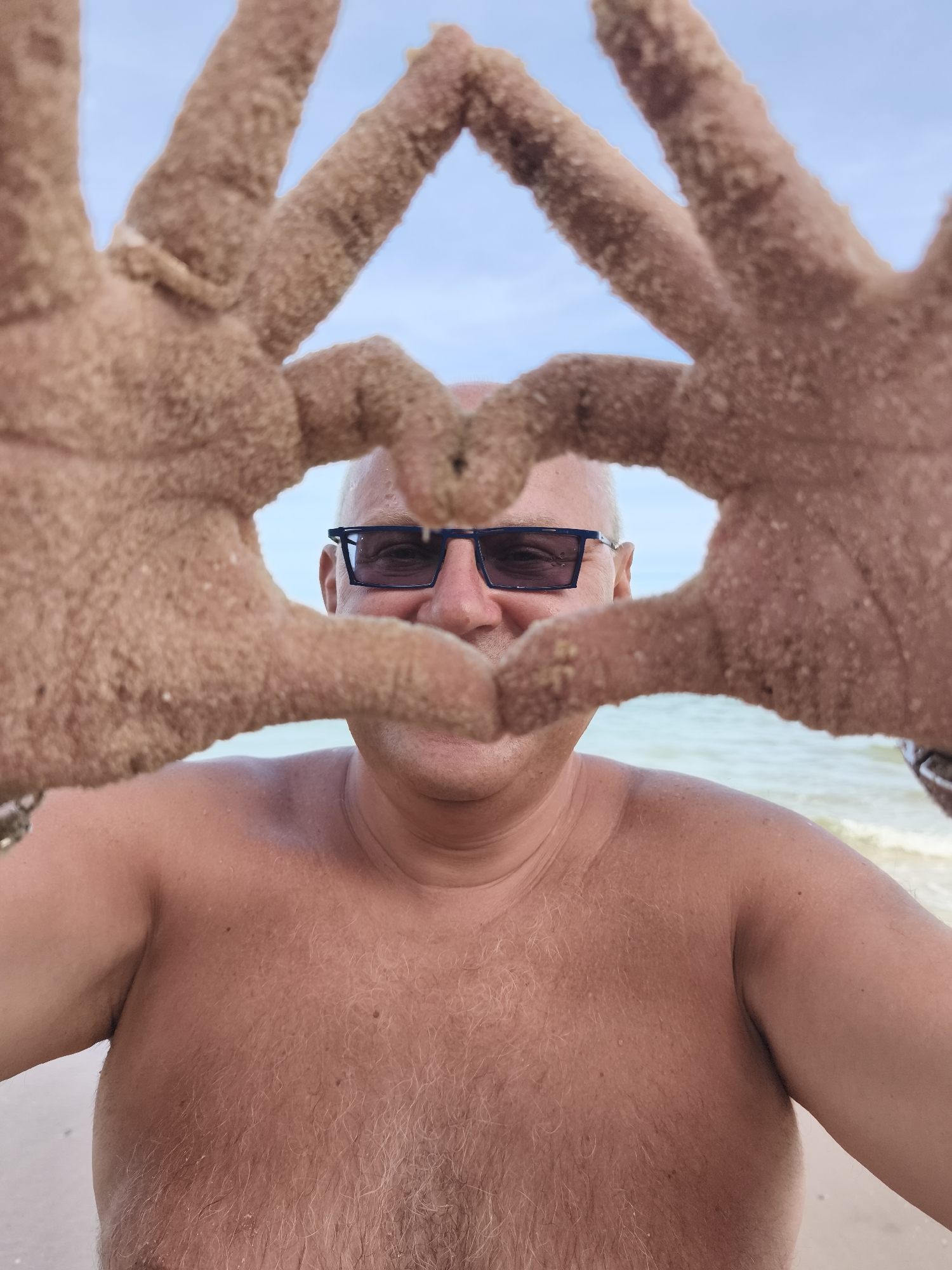 A middle-aged man, with fair skin and a bald head, is pictured at a beach. He is wearing dark-rimmed sunglasses and is shirtless, exposing his chest, which is covered in light-colored hair. His hands, covered in sand, are held up in front of his face, forming a heart shape. The background shows the ocean and a light blue sky. The location is identified as Phu Thuy Ward, Vietnam.