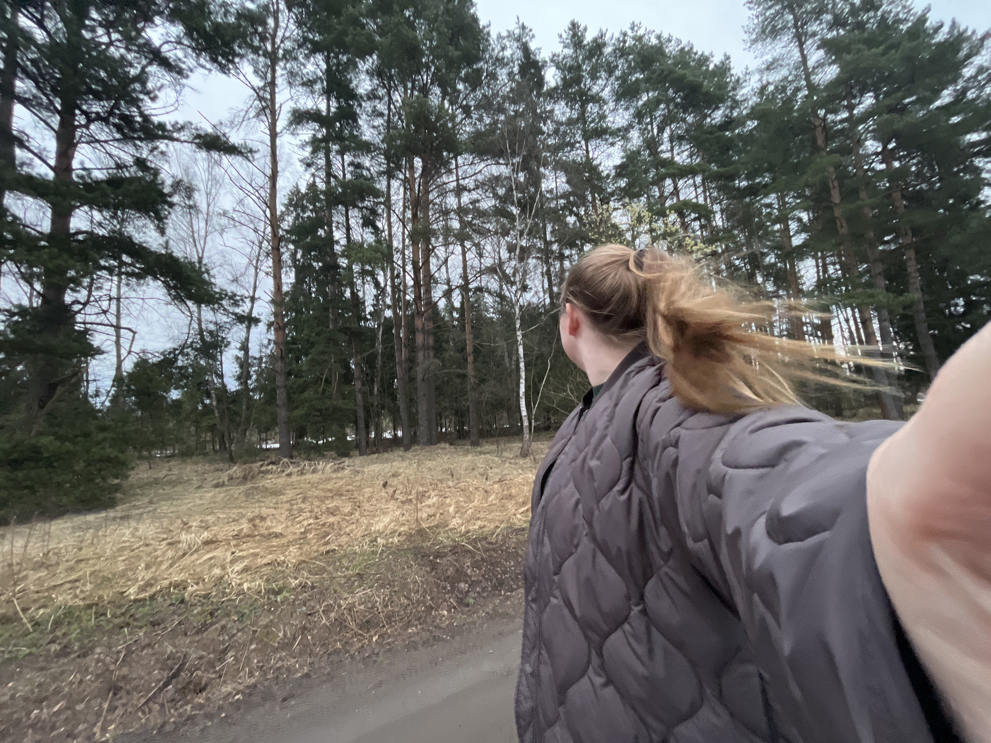 A person stands on a dirt road next to a field of dry grass and a forest of tall trees. The person is wearing a quilted grey jacket and has their blonde hair pulled back in a ponytail. The photo is taken from a low angle, with the person's arm visible in the foreground. The trees in the background are tall and dense, with a few bare trees mixed in. The sky is overcast. The location is Razdolye, Russia.