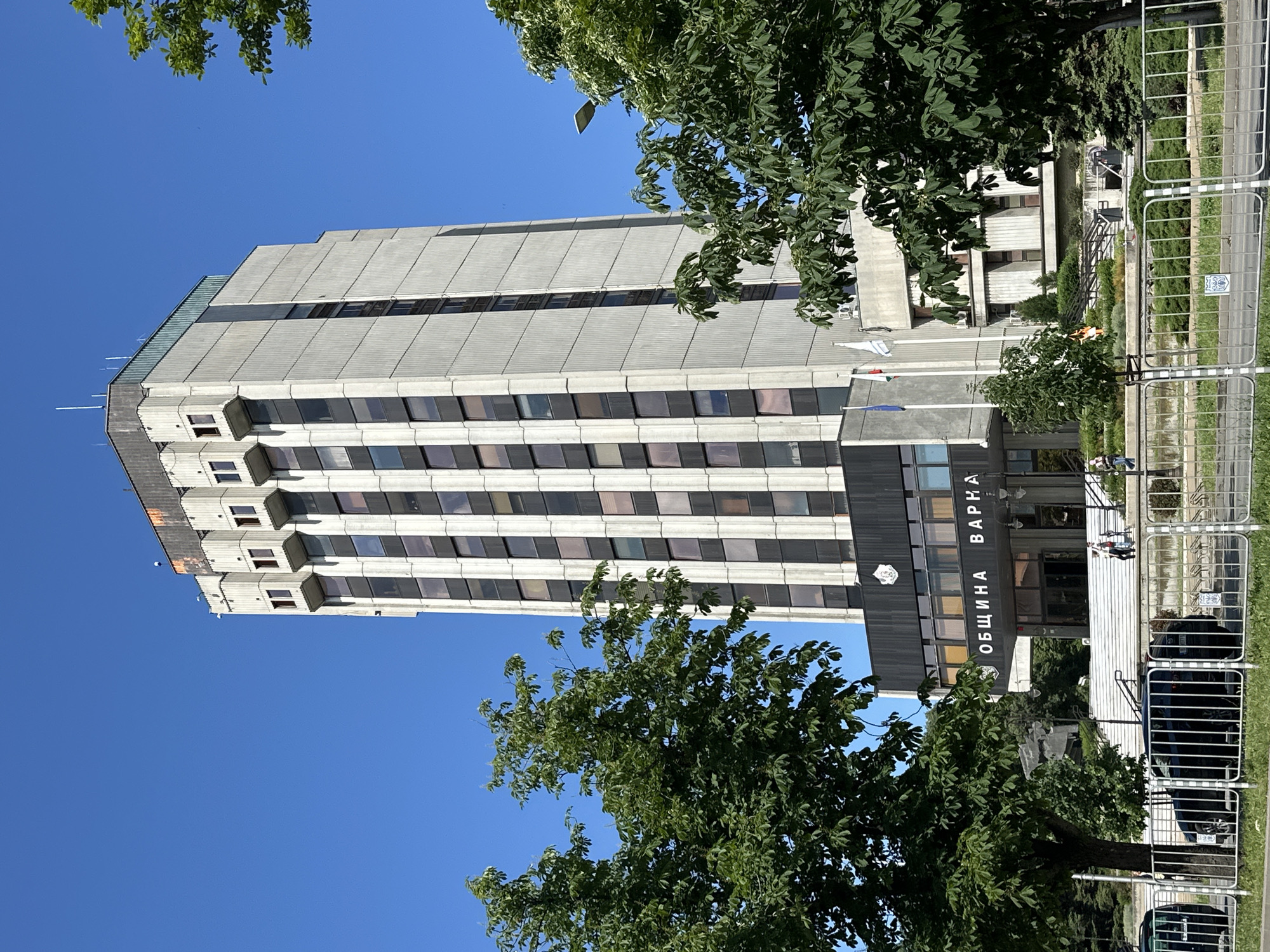 The image shows a tall, multi-story building with a predominantly grey facade and dark windows, partially obscured by trees. A sign in Cyrillic script (ОБЩИНА ВАРНА) indicates that the building is the Municipality of Varna (city hall) in Varna, Bulgaria. The building is constructed in a brutalist architectural style. A temporary pedestrian barricade surrounds the building's front plaza. The sky is clear and blue.