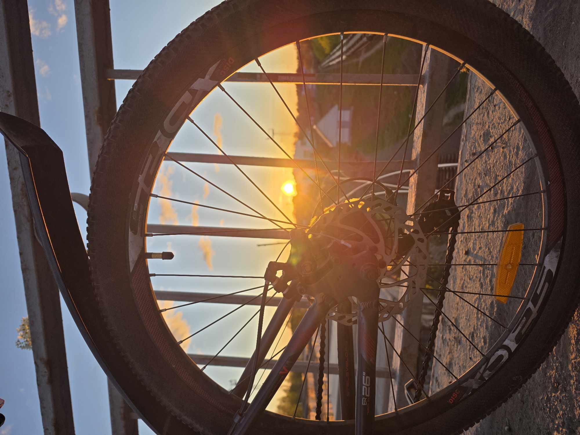 The image captures a close-up view of a bicycle wheel against a backdrop of a sunset in what is likely Chaikovsky, Russia. The wheel, with its spokes radiating from the hub, is framed by the setting sun, casting a warm glow on the scene. The tire's tread and sidewall markings are visible, along with the disc brake rotor. The background is blurred due to the shallow depth of field but appears to show buildings and infrastructure, indicative of an urban environment. The composition suggests the bicycle is positioned on or near a concrete structure. The lighting indicates that the photograph was taken during the evening hours.