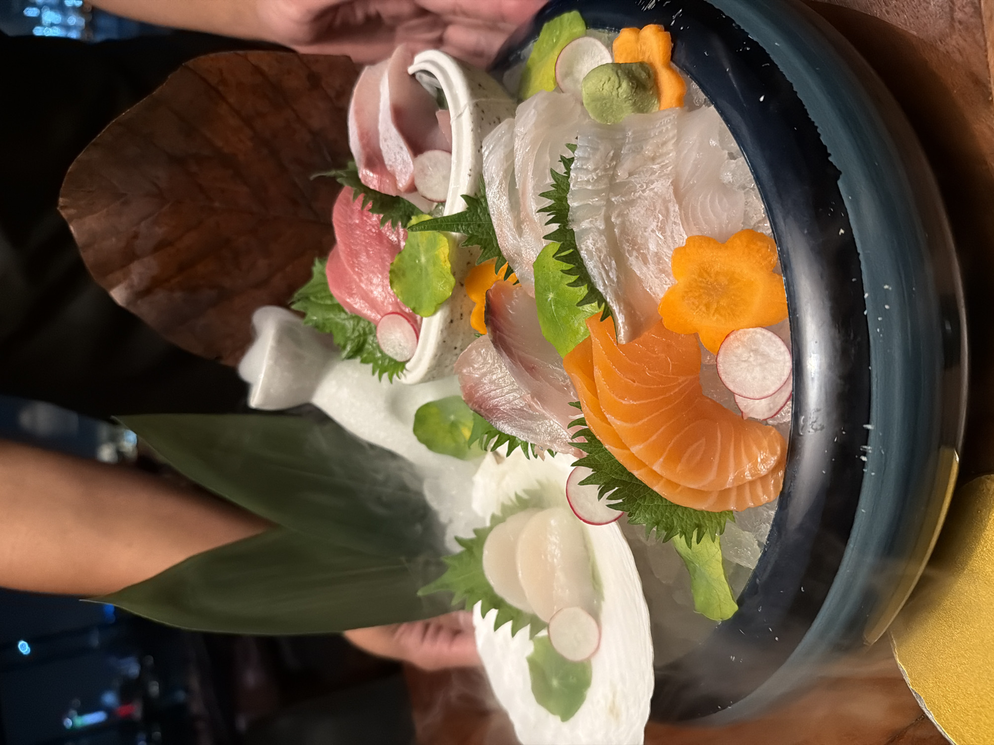 The image shows a close-up, eye-level view of an elaborate sashimi platter served on a dark blue, wide-rimmed ceramic bowl filled with crushed ice. The platter is presented with dramatic steam rising from the ice, creating a theatrical effect. The sashimi includes vibrant slices of salmon, several types of white fish, and pieces of tuna and lighter colored fish arranged in a decorative manner. Garnishes include finely cut radish slices, shiso leaves, carved carrots shaped like flowers, and a small mound of wasabi. Two large green bamboo leaves frame the left side of the platter, and a single, large, dried brown leaf is positioned behind the arrangement. A person's arm and hands are visible in the background, holding the platter or presenting it. The background is blurred, showing a dark, ambient setting with hints of city lights, suggesting an upscale restaurant or bar environment, potentially in Dubai, United Arab Emirates.