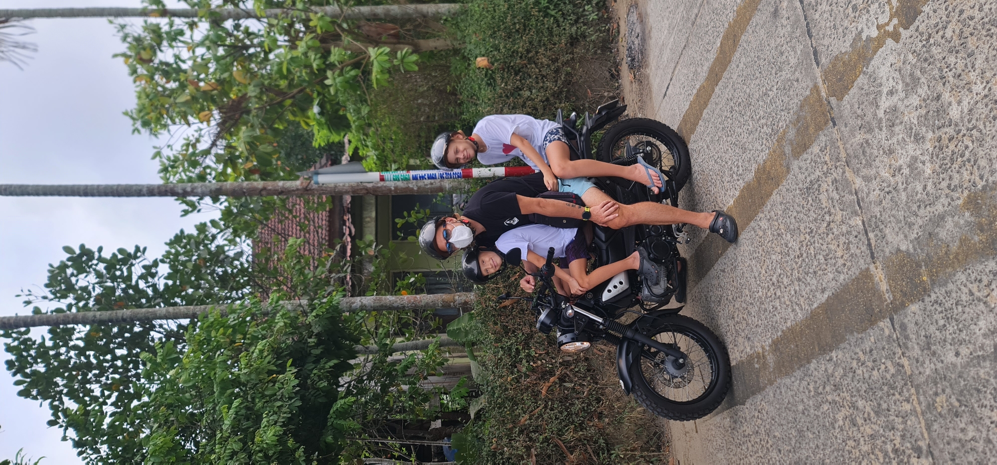A medium outdoor shot depicts three people on a black motorcycle on a paved road in the Hội An Ward, Vietnam. An adult wearing a black t-shirt, helmet, sunglasses, and a white face mask sits in the motorcycle's driver seat. A child with a helmet and a white t-shirt with a red heart design is behind the driver. Another child with a helmet and a white t-shirt sits in front of the driver on the motorcycle's seat. The road is gray concrete with faded yellow stripes. Lush green foliage flanks the road. Buildings and utility poles are visible behind the foliage. The sky is overcast.