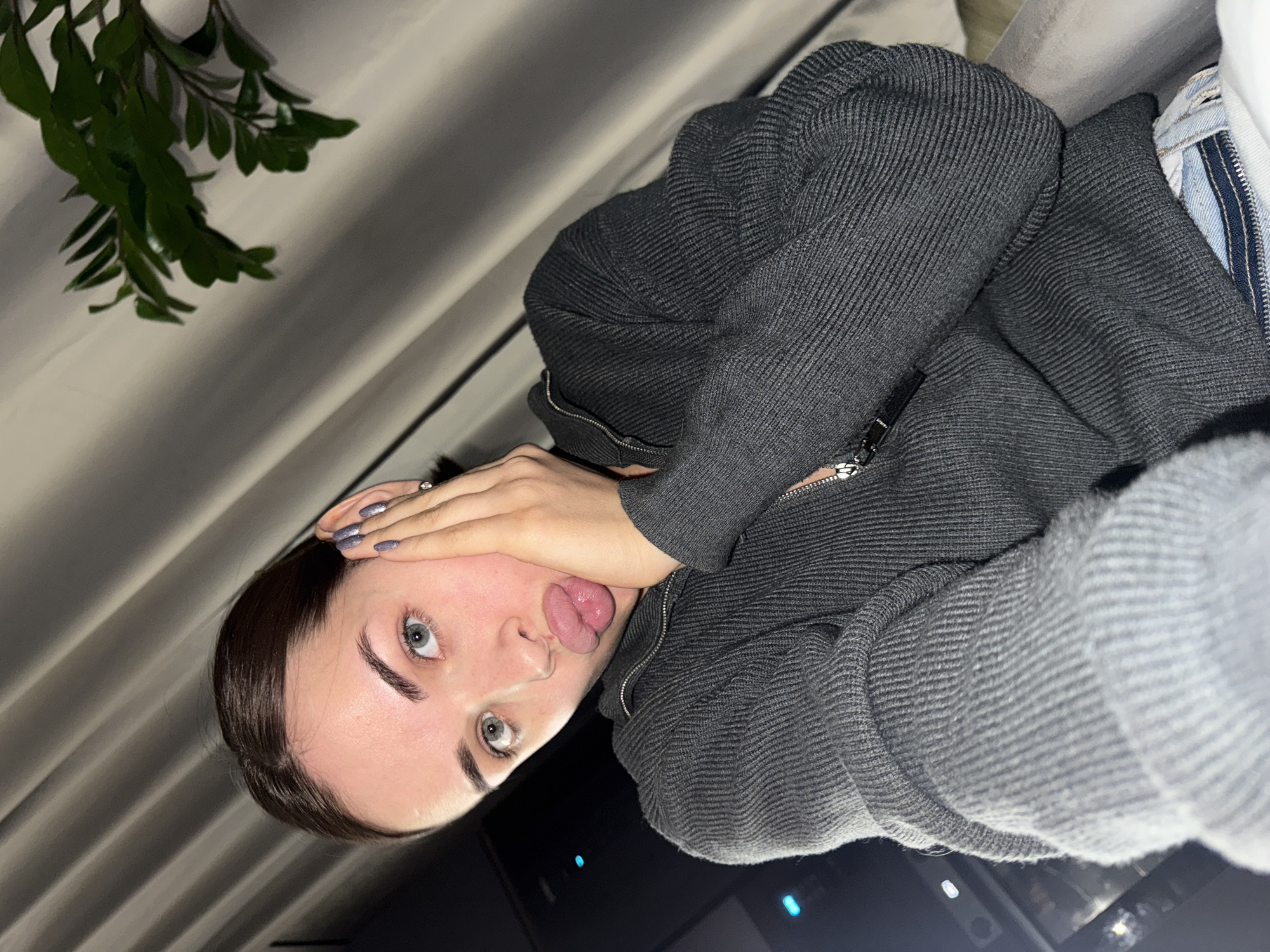 A young woman with fair skin, dark eyebrows, and light eyes is facing the camera. Her head is tilted to the left. Her hand is raised to her forehead, and she is sticking her tongue out. She is wearing a dark gray, zippered sweater. In the background are light-colored curtains and green foliage from a plant. The location is uncertain and unrelated to Лобаново, Russia.