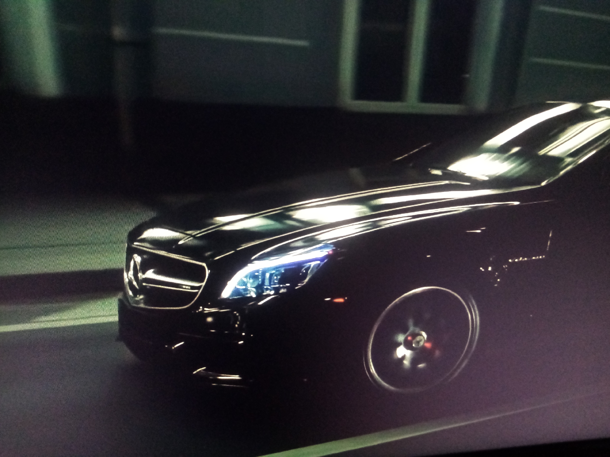 The image shows the front of a black Mercedes-Benz sedan, possibly a CLS model, at night. The car is moving, as indicated by the motion blur on the front wheel, which has a red brake caliper visible. The headlights are on, emitting a bright blueish-white light, and the car's grille features the Mercedes-Benz logo prominently. The scene appears to be in an urban or suburban area, possibly a street or a parking lot, with a dark background that suggests buildings or structures with illuminated windows. The lighting is dramatic, with strong highlights on the car's polished surface and reflections on the road. The overall mood is sleek and sophisticated. No people are visible in the frame. No visible text.