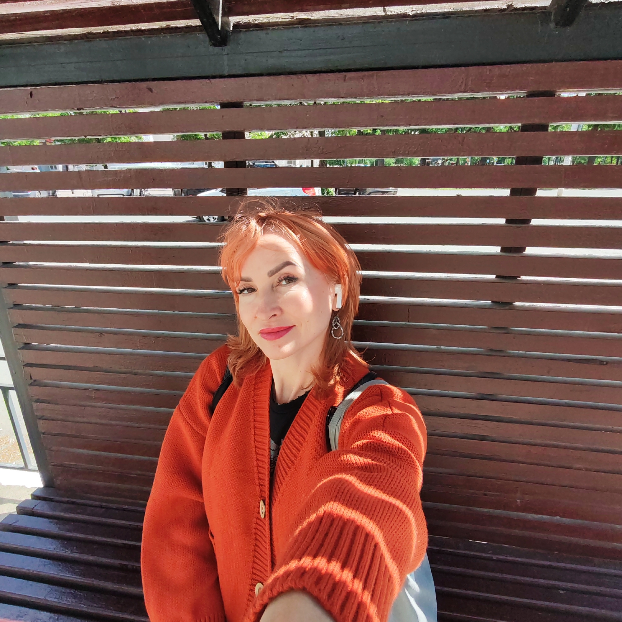 A woman with red hair is sitting on a wooden bench in what appears to be an outdoor public space. She is wearing an orange cardigan and has an earpiece in her left ear. Behind her is a wooden structure, possibly a bus stop or pergola, with horizontal slats that allow a view of the street and trees in the background. The photo is taken from a close-up perspective, suggesting it may be a selfie. The location is identified as the city of Chaikovsky, Russia, but the specifics of that location are not visible in the image.