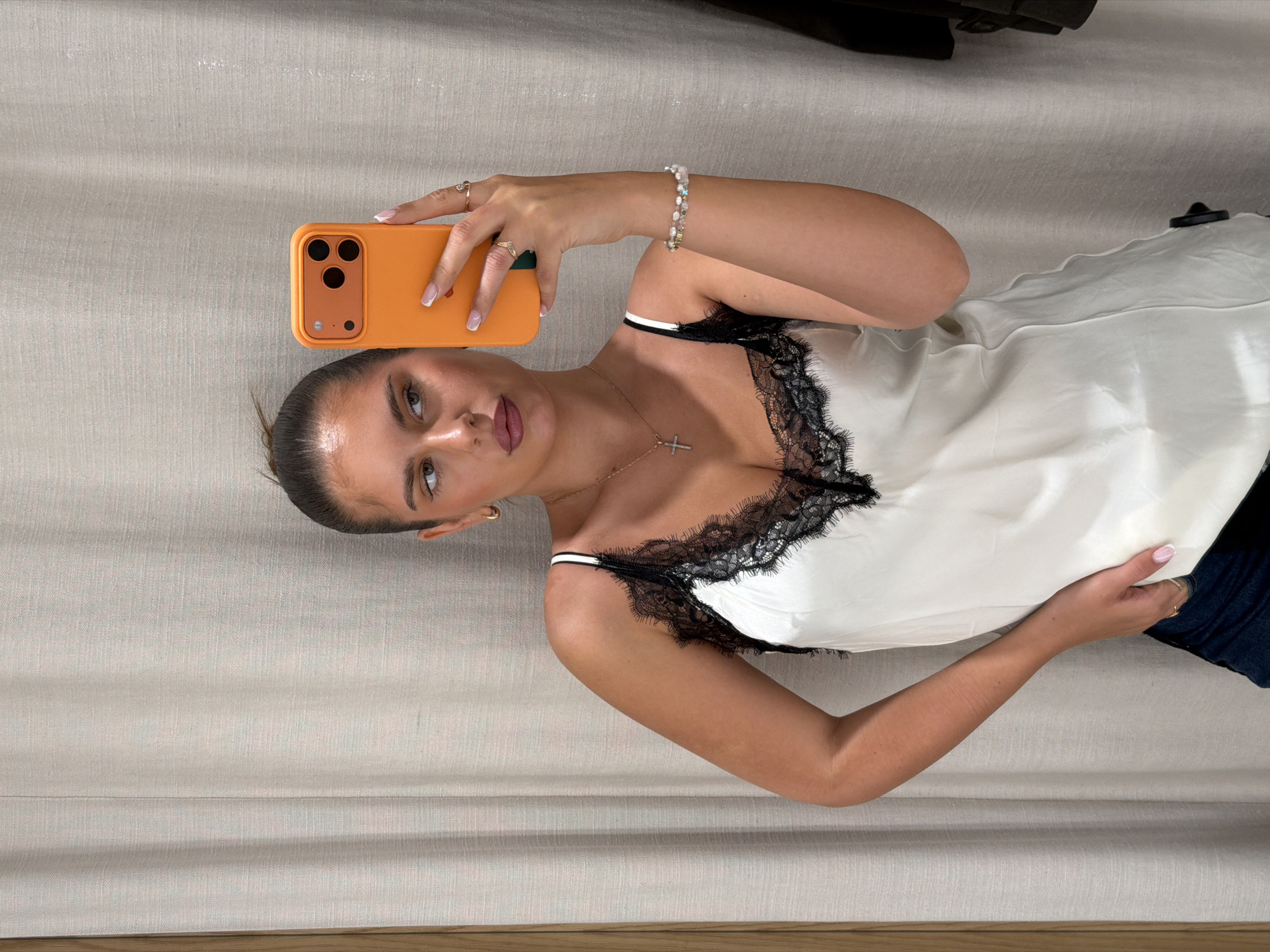 A woman is taking a selfie with an orange mobile phone. She is wearing a white satin tank top with black lace trim and a gold necklace with a cross pendant. She is also wearing a bracelet. The background appears to be a neutral-colored couch or bed. The location is not discernable to be the city of Krasnoyarsk, Russia.