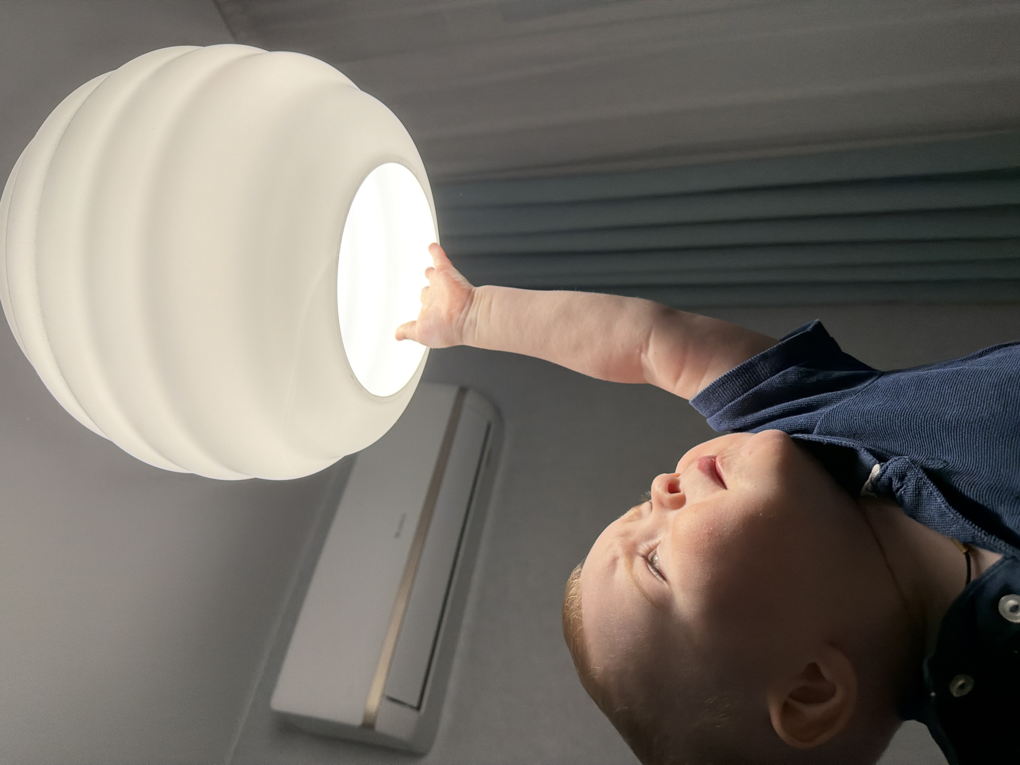 A baby in a navy blue polo shirt reaches up towards a large, round, white ceiling light. The background features a light gray wall and a white air conditioning unit with silver trim. The scene's location in Votkinsk, Russia, cannot be determined from the visual information provided.