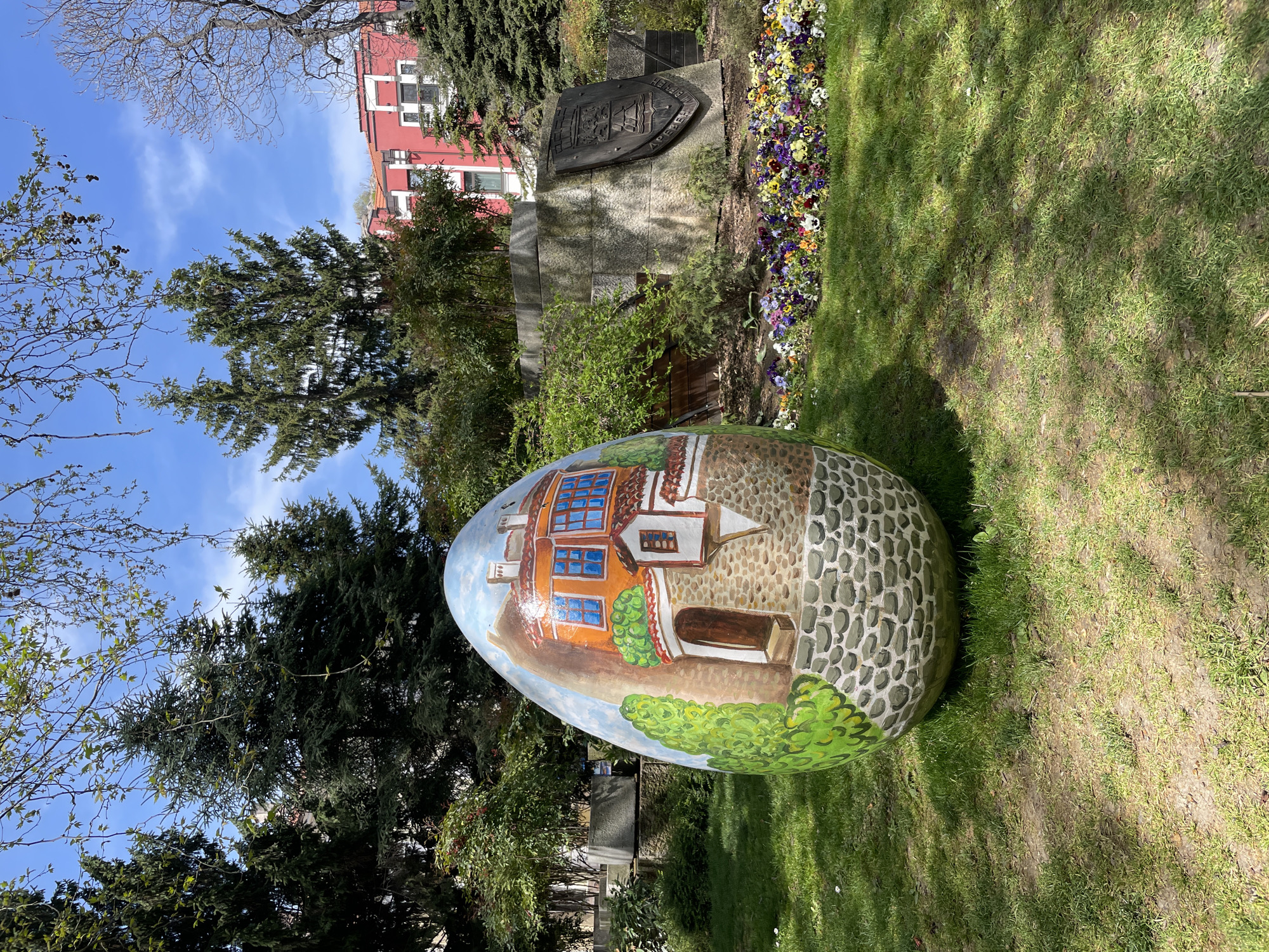 A large, intricately painted egg-shaped sculpture is the central focus of this outdoor scene, set in a park-like environment. The sculpture depicts a charming, multi-storied house with orange-brown walls, distinct blue-framed windows, and a lower section rendered in a traditional stone pattern, alongside green bushes and a sky motif.
The egg rests on vibrant green grass, which shows shadows indicating bright sunlight. In the mid-ground, behind the egg, a stone monument or plaque is visible. It features an embossed crest or emblem and Cyrillic text, which upon closer inspection, appears to include "ПЛОВДИВ" (Plovdiv) and "ДРЕВЕН" (Ancient), referring to the ancient city. A colorful bed of pansies, displaying hues of purple, yellow, and white, blossoms beside the monument.
The background features a clear blue sky with a few faint clouds. Various trees, both dark green coniferous types and deciduous trees with budding leaves, frame the upper portion of the image. A red building with white window frames can be seen behind the trees and monument.
The scene captures a peaceful, sunny daytime moment, showcasing public art and landscaping. The visible text on the monument, combined with the architectural style depicted on the egg, firmly places the location in Plovdiv, Bulgaria. There are no people or specific activities taking place, just a serene landscape with artistic elements.