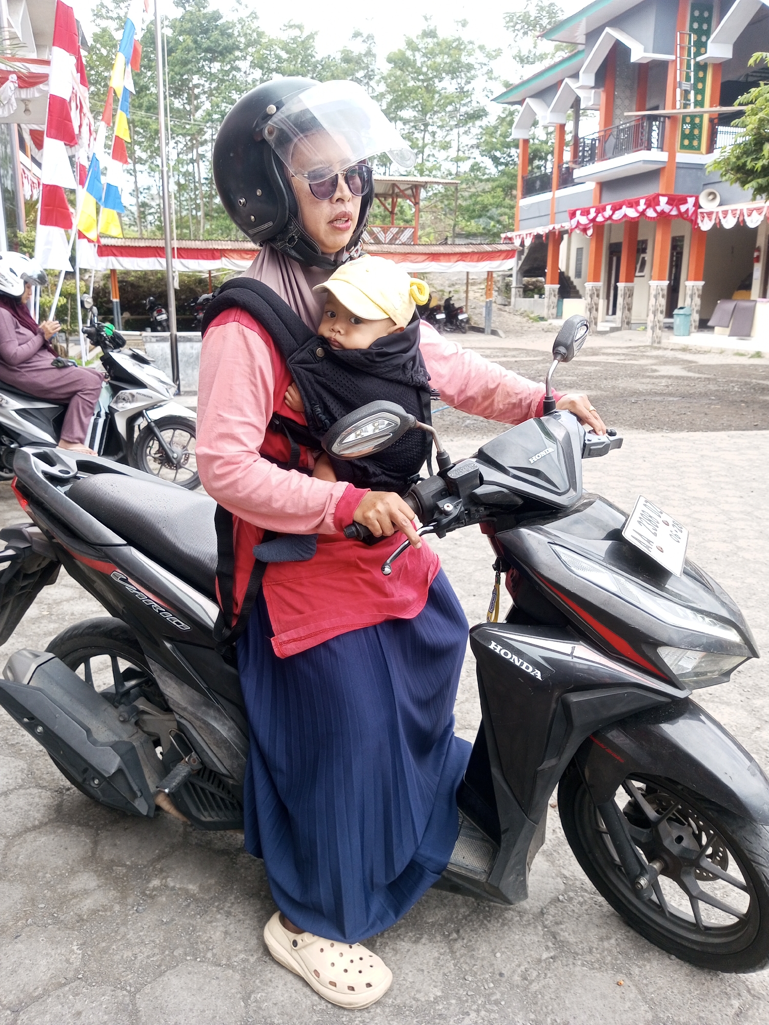 A woman wearing a black helmet with a clear visor, sunglasses, and a pink long-sleeved shirt over a black baby carrier, is seated on a black Honda motorcycle. A baby wearing a yellow hat is nestled in the carrier. The woman wears a dark blue pleated skirt and cream-colored slip-on shoes. Another woman wearing a purple headscarf and matching outfit is seated on a white motorcycle in the background. The motorcycles are parked on a paved surface in front of a building with an orange and gray facade, adorned with red and white banners. Several flags are visible on poles to the left of the building. The environment appears to be outdoors in a village or town setting, possibly in Krasak, Indonesia, given the presence of the motorcycles and the architectural style. The overall scene depicts a casual moment of transport or waiting.