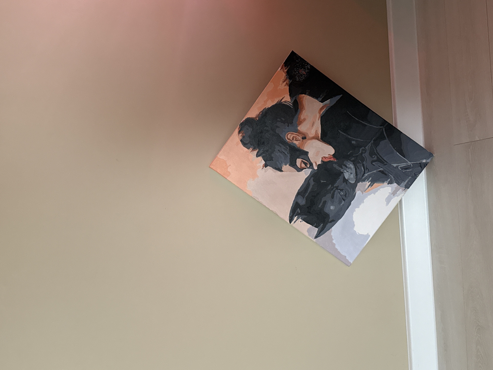 A painting of Batman and Catwoman kissing is hung on a light beige wall next to a white door frame and light wood paneled door in a room of Unknown, Unknown. The painting is a square and is tilted at an angle.