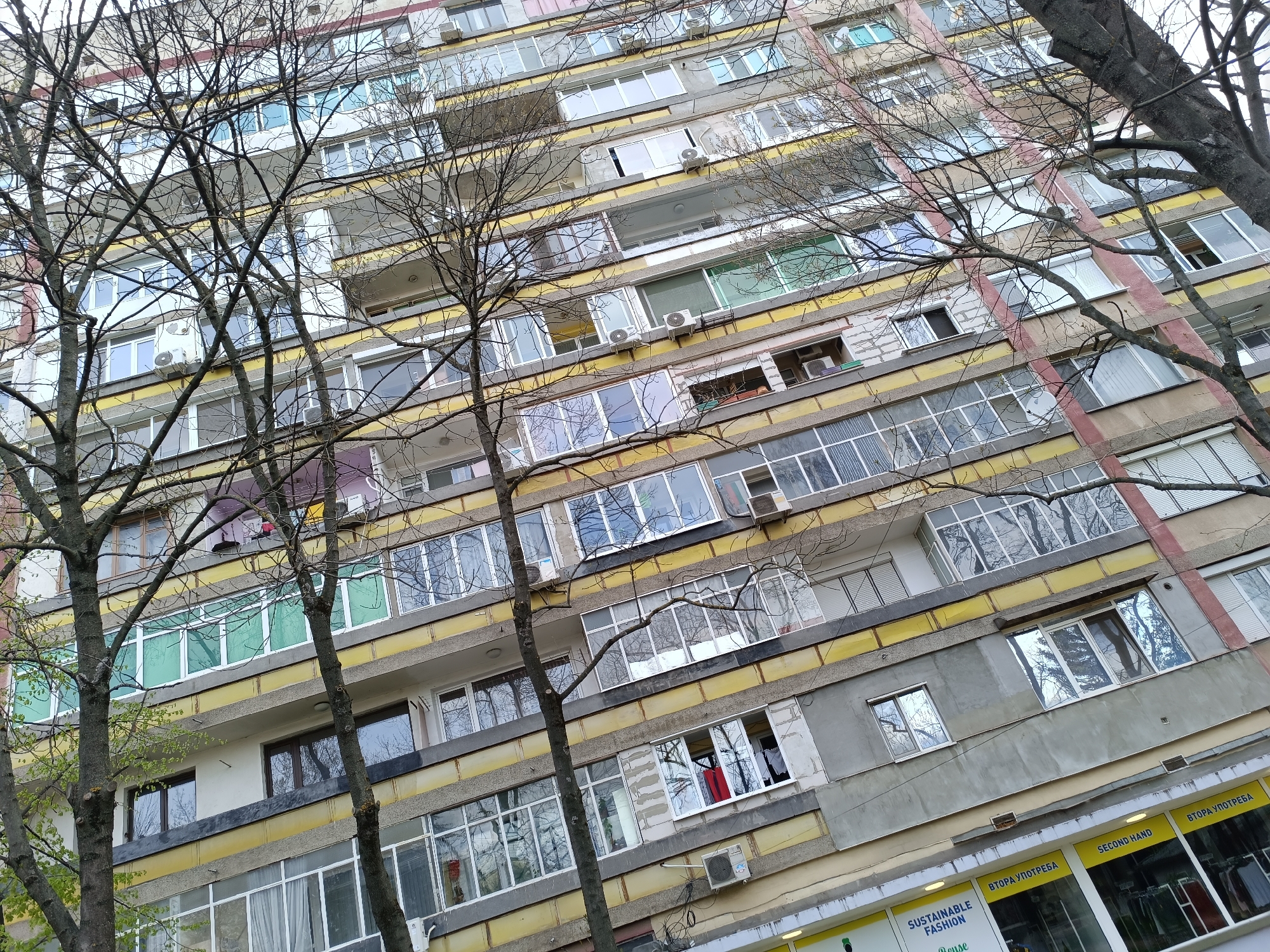 A low-angle shot shows the facade of a multi-story building, framed by trees with bare branches. The building has a repetitive pattern of windows, air conditioning units, and balconies, painted in alternating bands of yellow and gray. At the bottom of the image, storefront signs with the words "SUSTAINABLE FASHION" and "SECOND HAND" can be seen. The sky is overcast, and the overall impression is that of a typical urban setting. The location is in the city of Unknown, Unknown.