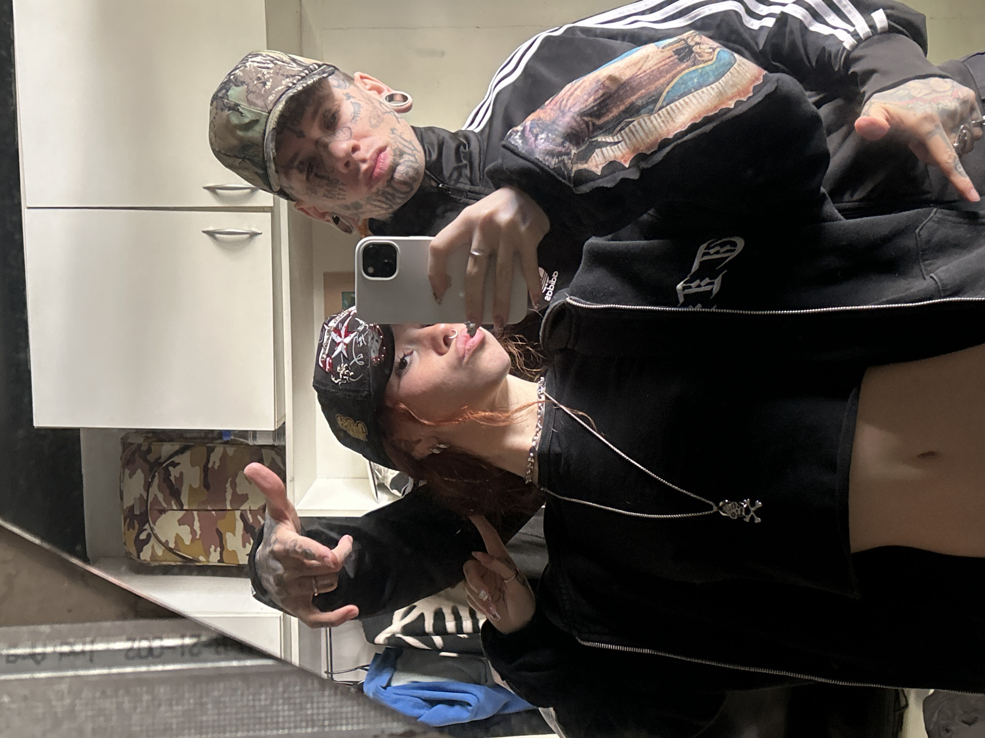 Two individuals, a man and a woman, are captured in a mirror selfie in an indoor setting, possibly a bathroom or hallway, in Buenos Aires, Argentina. The image is rotated, with the ceiling appearing on the right.
The man, positioned in the upper portion of the frame, has extensive facial tattoos, ear gauges, and is wearing a camouflage baseball cap. He is dressed in a black Adidas track jacket with white stripes, featuring a prominent tattoo of religious iconography (likely Our Lady of Guadalupe) on his right arm sleeve. He holds a white smartphone, presumably an iPhone, to take the photo. His left hand, also tattooed and adorned with rings, is visible, pointing downwards with his thumb extended.
The woman, in the lower part of the frame, has reddish-brown hair and is wearing a black baseball cap with an embroidered design, a black cropped jacket, and a silver chain necklace with a skull and crossbones pendant. A nose piercing is visible, and she has tattoos and rings on her fingers. Her midriff is exposed below her jacket. She poses with a slight pout and her right hand also shows tattoos and rings.
In the background, white cabinets with silver handles are visible. An open cabinet reveals a camouflage-patterned bag or box. The reflective surface in the foreground shows distorted reflections, including what appears to be a metal grate or textured floor, with reversed partial text like "200-12." The "adidas" logo is visible in reverse ("ebibba") on the man's jacket. The overall atmosphere is casual and contemporary, reflecting a personal moment.