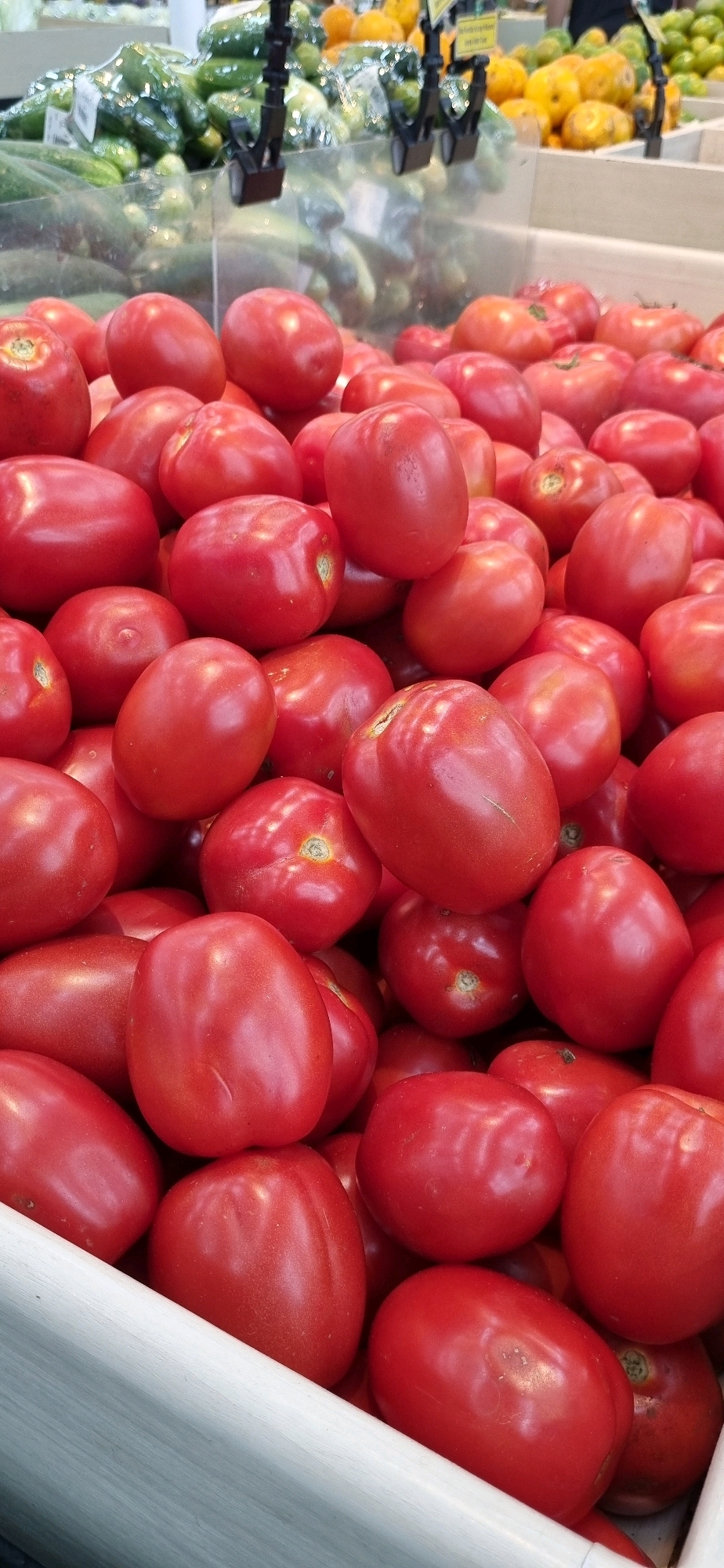 A close-up, overhead shot features a vibrant display of ripe, red Roma tomatoes piled high in a white wooden crate. The tomatoes are a rich crimson color, with a smooth, slightly glossy skin. Some are plump and oval-shaped, while others appear more elongated. The crate itself is made of light-colored wood with visible grain. In the blurred background, a display of cucumbers and what appear to be yellow squash or lemons can be seen, suggesting this scene is from a grocery store or market in Denpasar, Indonesia, likely within a produce section. The lighting is bright, illuminating the fruits and casting soft shadows. There are no people visible in the image.