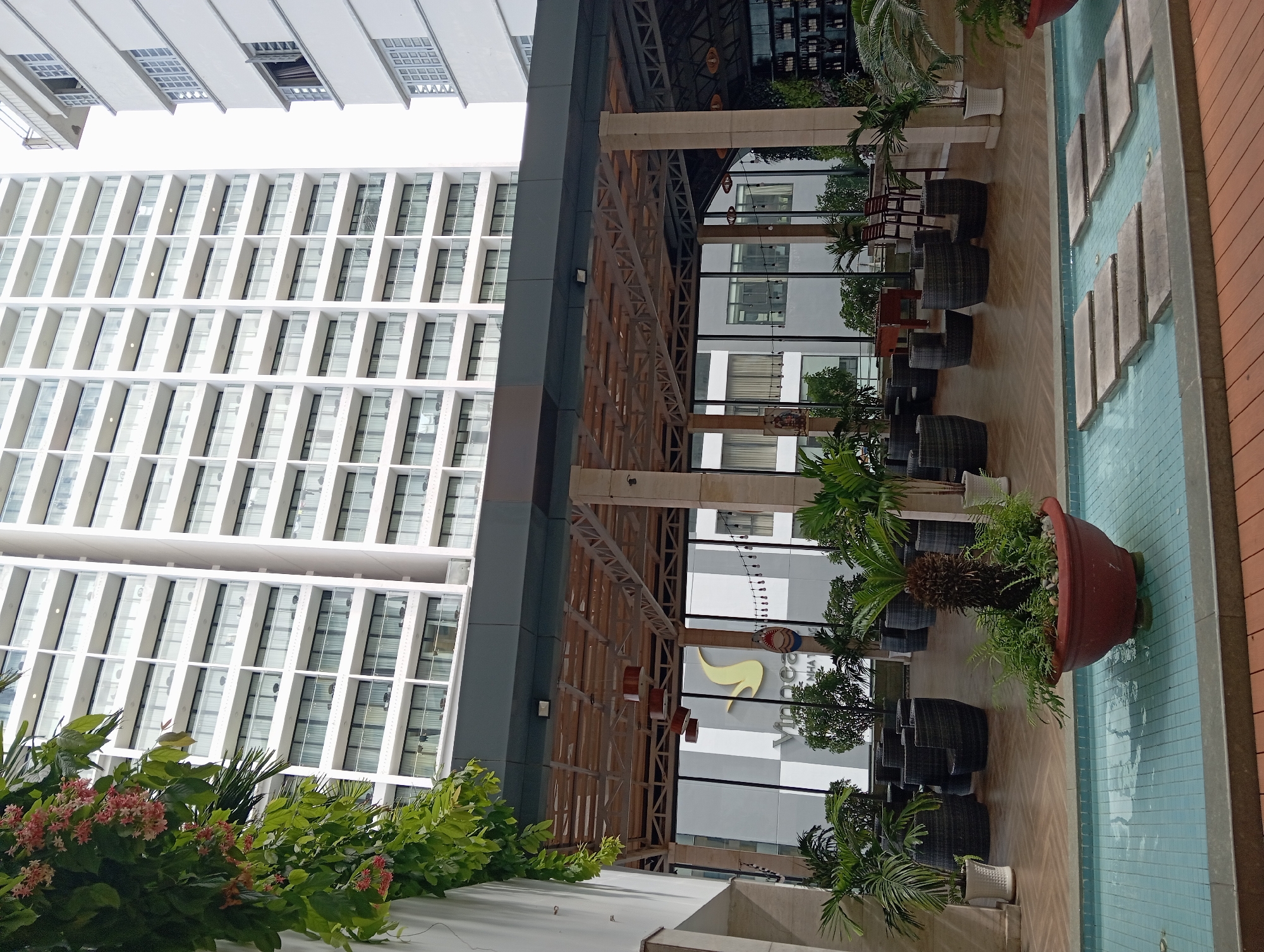 This image shows an outdoor dining or lounge area in Nha Trang, Vietnam, situated between a body of water and a tall, modern building. The foreground features a shallow, tiled pool with stepping stones leading across it. A large, red planter with greenery sits in the pool. The midground is an open-air covered patio with a wooden slatted roof supported by pillars. It is furnished with dark wicker chairs and tables, arranged in a conversational setting. Beyond the patio, through large glass windows, a building with a distinctive yellow logo that appears to be for "Vinpearl Nha Trang" is visible. The background is dominated by a very tall, white building with numerous balconies and windows, characteristic of a hotel or residential high-rise. The sky is overcast. No people are visible in the scene.