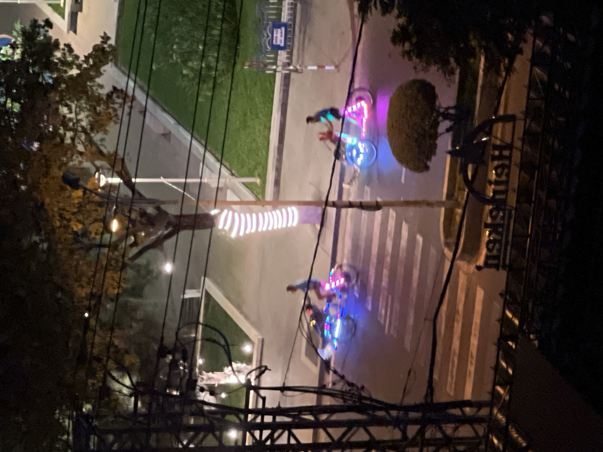 At night in Nha Trang, Vietnam, two cyclos, illuminated with colorful LED lights, travel along a street. One cyclo, in the foreground and on the right, is ridden by a man in a blue shirt, with a red-clad passenger behind him. The other cyclo, to the left, has a single passenger. The cyclos are crossing a pedestrian crosswalk. The street is lined with trees, one of which is adorned with white lights. In the background, a sidewalk and a grassy area are visible. A street lamp with a bright light stands on the left side of the image. A sign with the word "Heineken" is visible at the bottom of the image. The scene is captured from a slightly elevated vantage point, possibly a balcony or window, overlooking the street.