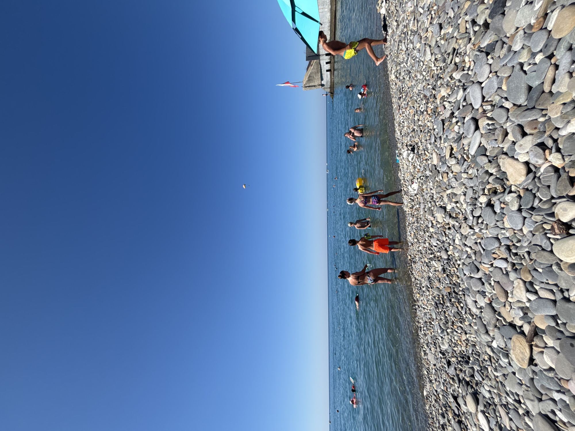 A wide shot shows a pebble beach and the sea under a clear blue sky. Several people are in the water, some swimming and others standing. On the beach, a few people are visible, including a person in yellow shorts walking towards the right of the frame, partially under a turquoise umbrella. In the background, a concrete structure with a red and white flag stands on a pier or breakwater. The sea is calm with small waves lapping the shore. The overall scene suggests a sunny day at a popular beach location.