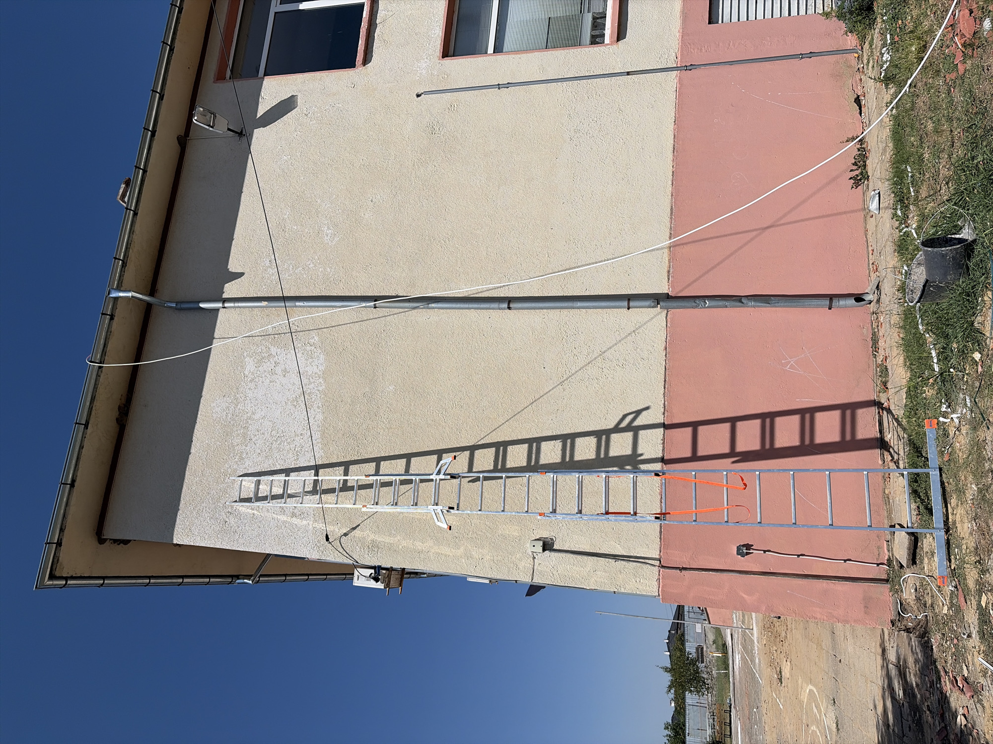 The image depicts an exterior view of a building located in Karageorgievo, Bulgaria. A tall, silver-colored ladder stands upright against the building's light beige and pink-painted facade. The ladder has orange straps and is positioned near a downspout. Various cables and wires are visible running along the building and on the ground. The ground around the building is unpaved and appears to be undergoing some form of construction or renovation, with debris, including bricks and white wiring, scattered on the ground. In the foreground, a black cylindrical object, possibly a bucket or pipe, lies on the grass. The sky above is clear and blue, indicating fair weather conditions. The building has visible windows and a gutter system.