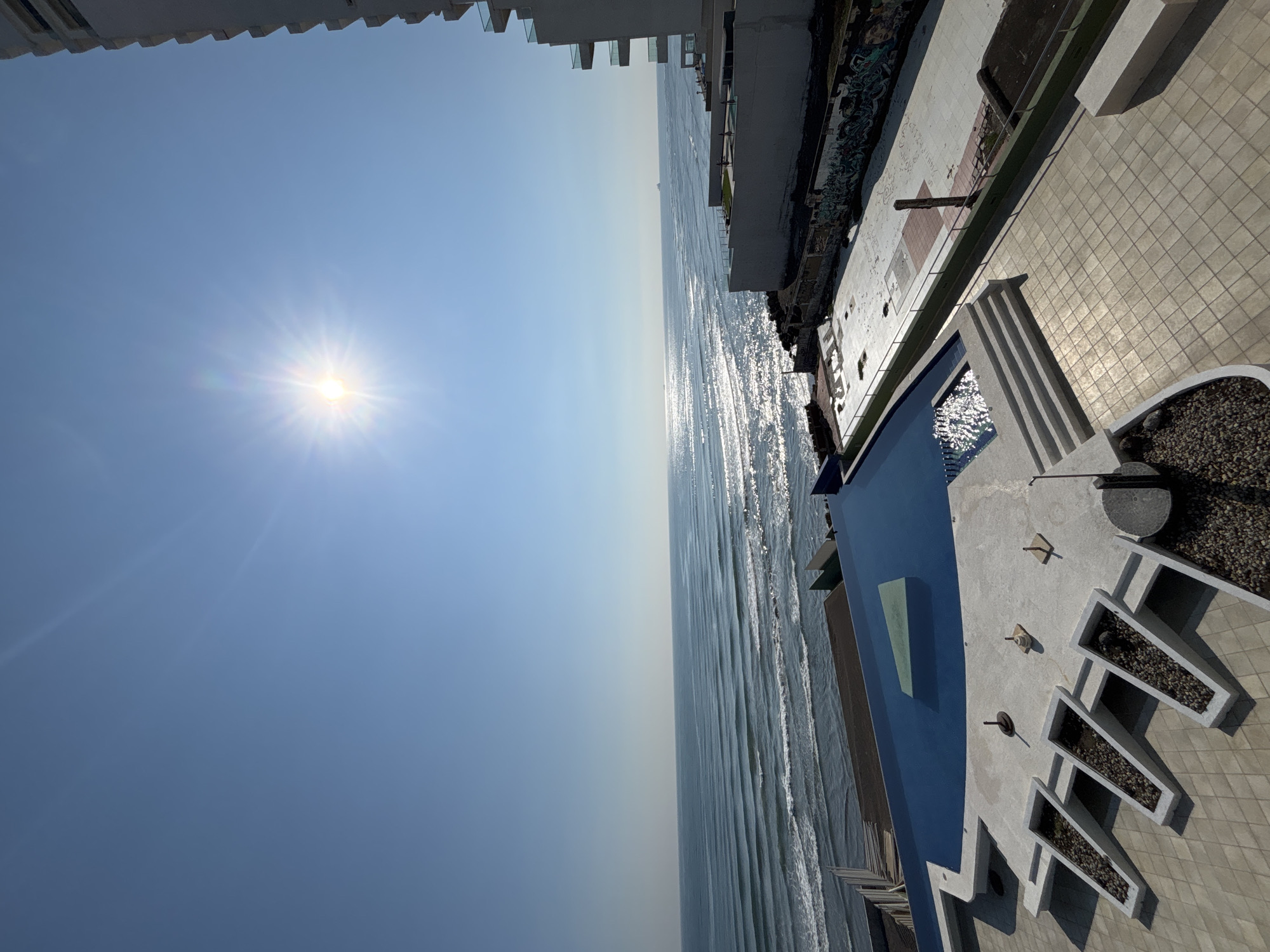 The image shows a high-angle view of the ocean, a building, and a pool, likely from a high-rise in Veracruz, Mexico. The ocean stretches across the middle of the frame, with sunlight reflecting on the waves. To the right, a building with a pool and patio area is visible. The sky is clear blue with the sun shining brightly. There are no people visible in the image.