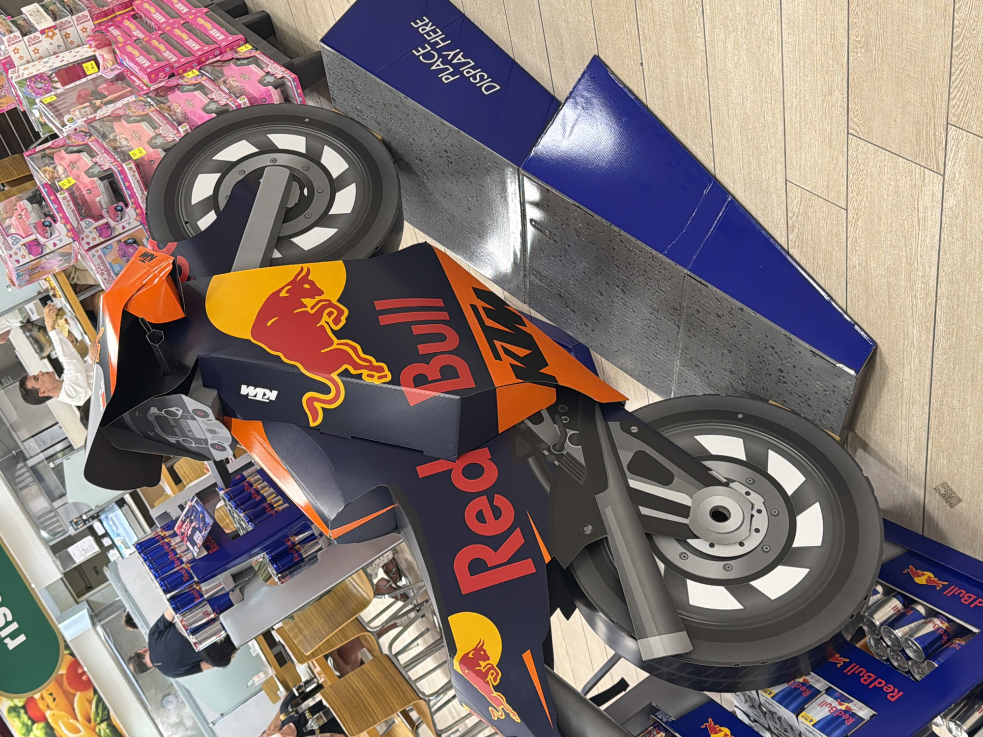The image captures a detailed view of a Red Bull branded KTM motorcycle display, likely in a supermarket or retail store environment. The display is constructed from cardboard and features the iconic Red Bull logo and KTM branding. The motorcycle is positioned on a stepped platform. In the background, several people are visible, some appearing to be customers or staff. One person is seen at a counter, possibly preparing food. Other individuals are seated at tables in what appears to be a dining area. Stacks of Red Bull cans are visible on shelves to the left of the display, and boxes of toys, possibly dollhouses, are stacked on shelves to the right. The overall setting suggests a commercial space with a variety of products and services available. The flooring is a light-colored wood-look laminate. The context of Due Carrare, Italy, indicates this scene is located within that specific town.