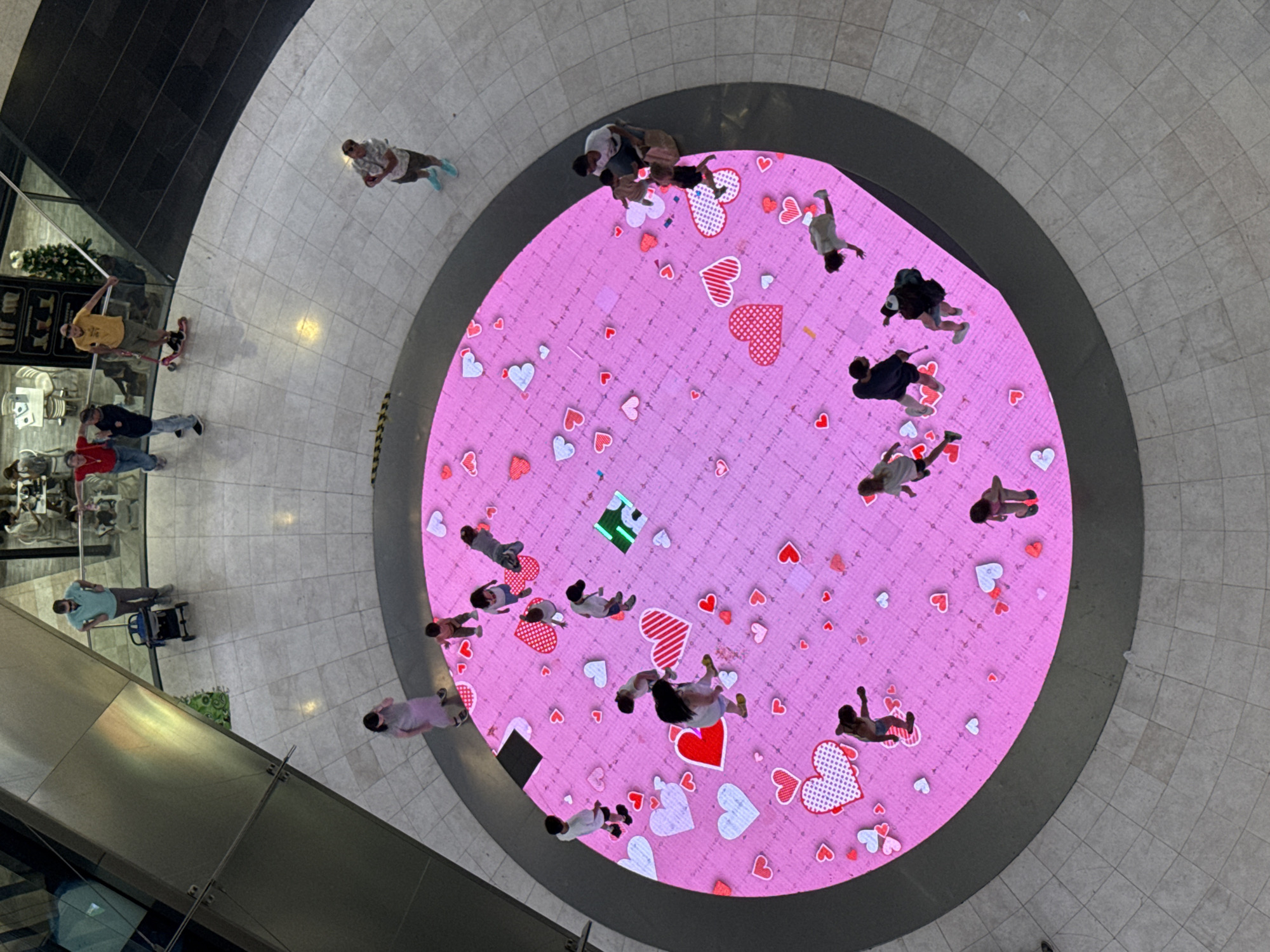 An overhead, aerial view captures a diverse group of people interacting with a large, circular, interactive LED floor display in a modern indoor public space, likely a shopping mall or atrium. The floor is predominantly covered with a vibrant pink background adorned with numerous red and white heart graphics of varying sizes, some with intricate patterns. Several individuals are actively engaged with the display, with some appearing to walk or stand on it, suggesting it's responsive to their presence.
In the background, outside the main circular display, the environment consists of polished beige tiled flooring and architectural elements indicative of a commercial setting. These include metal railings, glass partitions, and muted-toned walls. A few individuals are positioned around the perimeter of the display, observing or awaiting their turn to interact. One person on a scooter is visible further in the background, as is a decorated Christmas tree, indicating a possible festive season.
The people on and around the display are varied in age and attire. Their clothing ranges from casual wear to more athletic outfits. Some are alone, while others are in small groups. The overall atmosphere appears to be one of leisure and entertainment. The lighting is bright and even, typical of an indoor commercial space. The image quality is sharp, allowing for detailed observation of the individuals and the graphics on the floor. The location, as per the provided information, is the city of Barcelona, Spain, though the specific venue within Barcelona is not explicitly identified in the visual data.