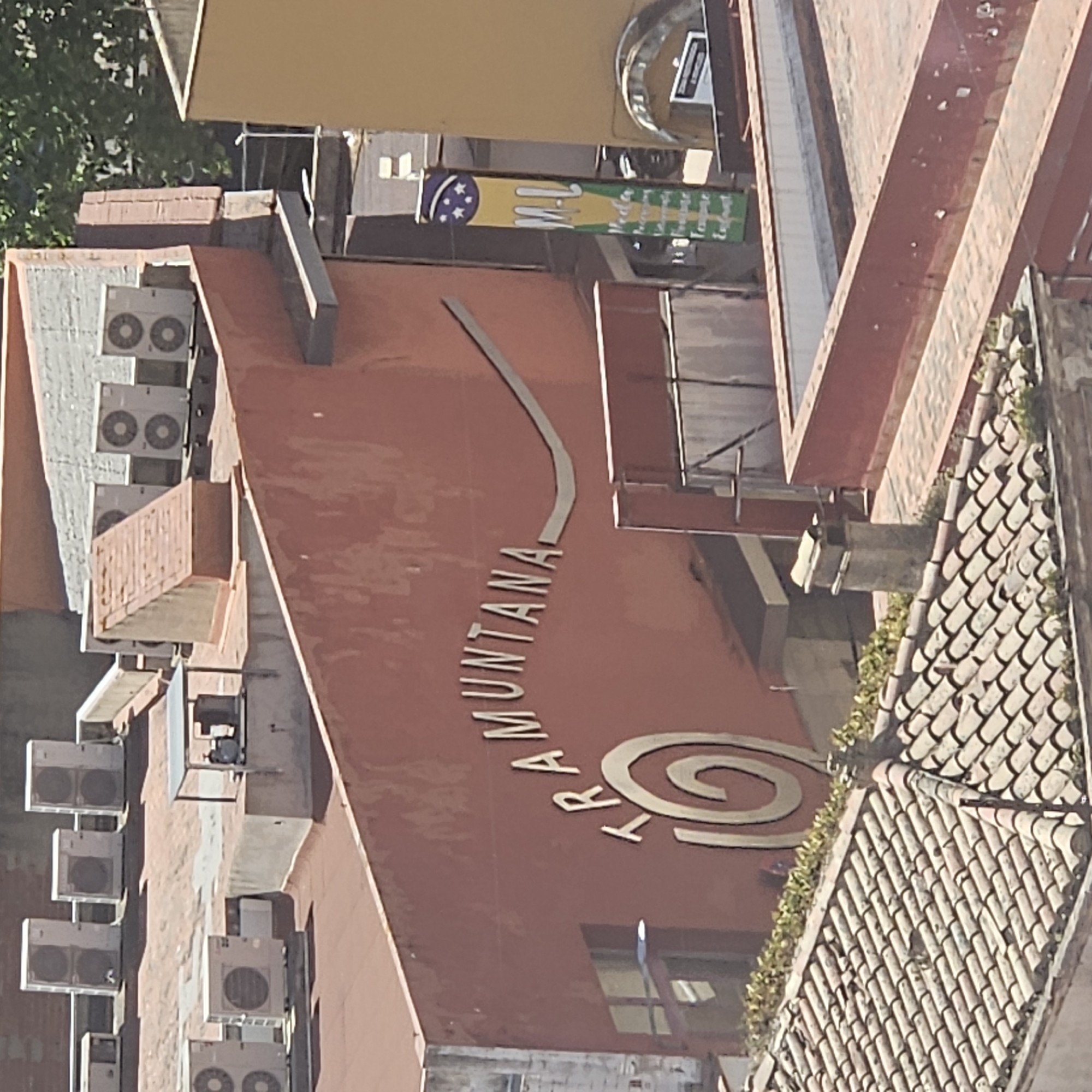 This is an exterior shot of a building in La Jonquera, Spain. A salmon-colored wall displays the word "TRAMUNTANA" in silver lettering. Near the beginning of the word is a silver spiral design. Air conditioning units are visible on the building's facade. Above the word Tramuntana, there is a banner with what appear to be flags from Brazil, with some lettering in green and yellow. To the right is a brown-tiled roof with some plant life. The overall angle of the image appears to be from a slightly elevated position.