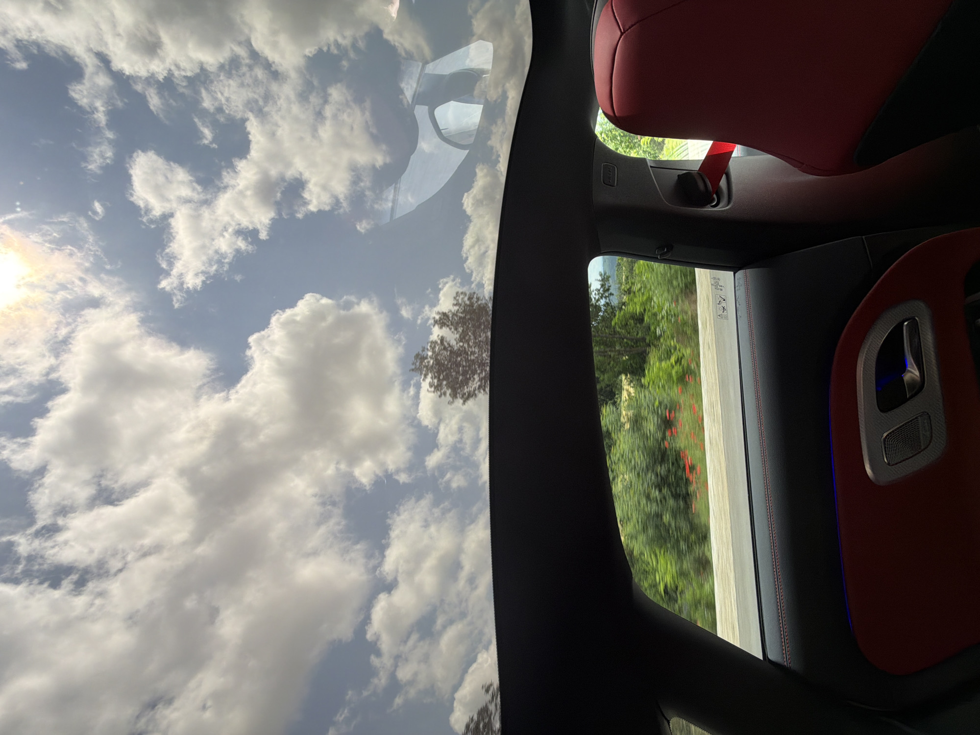 The image provides an interior view from a car, rotated 90 degrees to the left, looking outwards and upwards. When correctly oriented, the top of the image shows a bright blue sky filled with numerous white, fluffy cumulus clouds. The sun is visible in the upper left corner, partially obscured by clouds, casting a bright glare.
A large panoramic sunroof reveals the expansive sky. A faint, stylized reflection of the car's interior components is visible on the sunroof's surface.
To the right, the interior of the car is detailed. A plush red leather car seat, likely a headrest, occupies the upper right portion. Below it, a red seatbelt is fastened, with an "AIRBAG" label visible on the dark interior pillar. Further down, the car's door panel is visible, featuring dark upholstery with contrasting red stitching. A sleek silver handle or speaker grille is present on the door, accented by a subtle blue ambient light.
Through the side window, outside the dark car frame, there is a blur of vibrant green foliage, including trees and bushes, indicating the car is in motion. A light-colored road or barrier can also be glimpsed.
The scene takes place during a bright, sunny day, with clear weather and scattered clouds. The lush green vegetation is consistent with a natural or rural setting, potentially matching the provided location context of Sadovets, Bulgaria. No people are visible. The atmosphere suggests a pleasant journey.